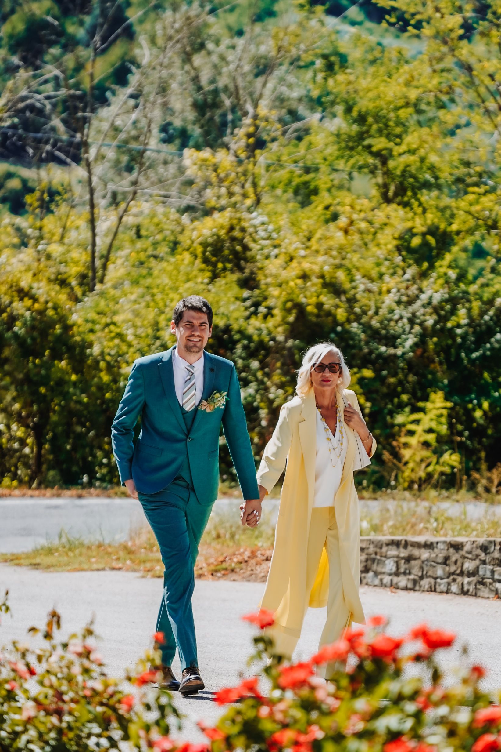 A groom in a teal suit and bride in yellow walk hand-in-hand outdoors with lush green trees and red roses.