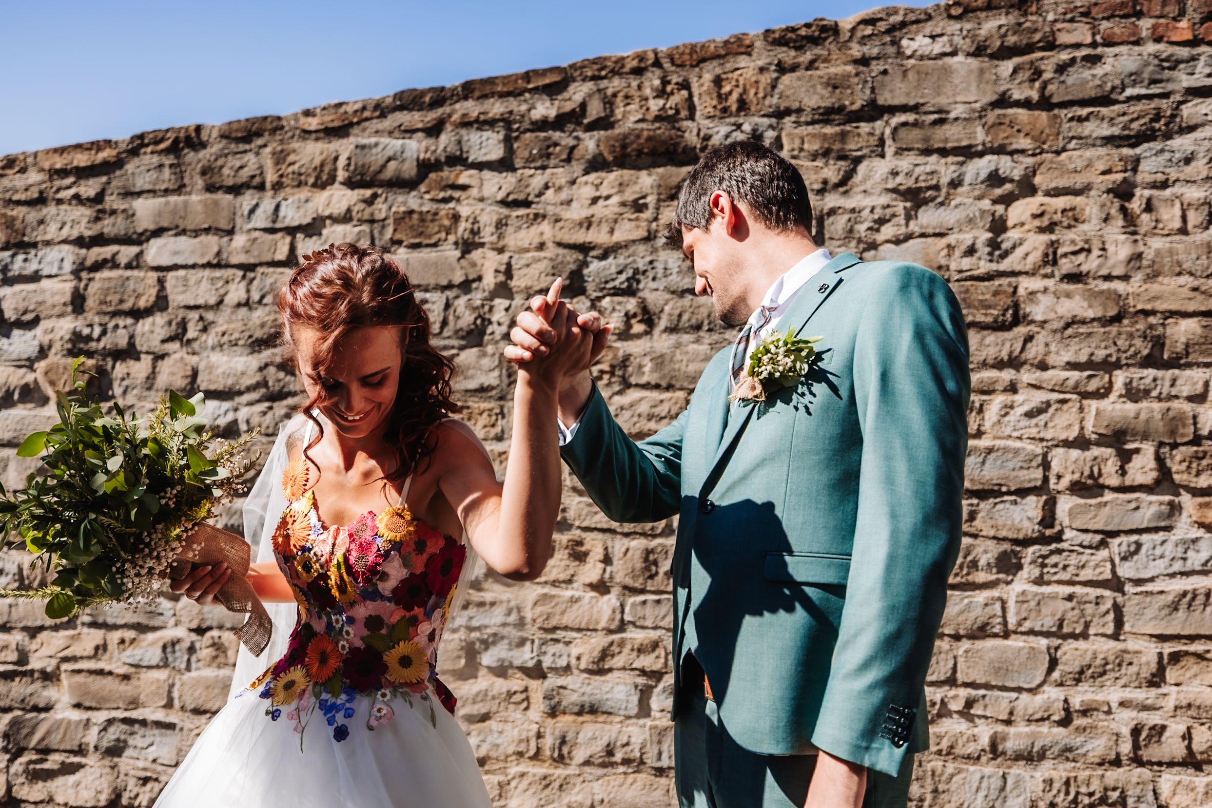 Bride and groom dancing outdoors against a stone wall, bride holding colorful bouquet in a white dress.