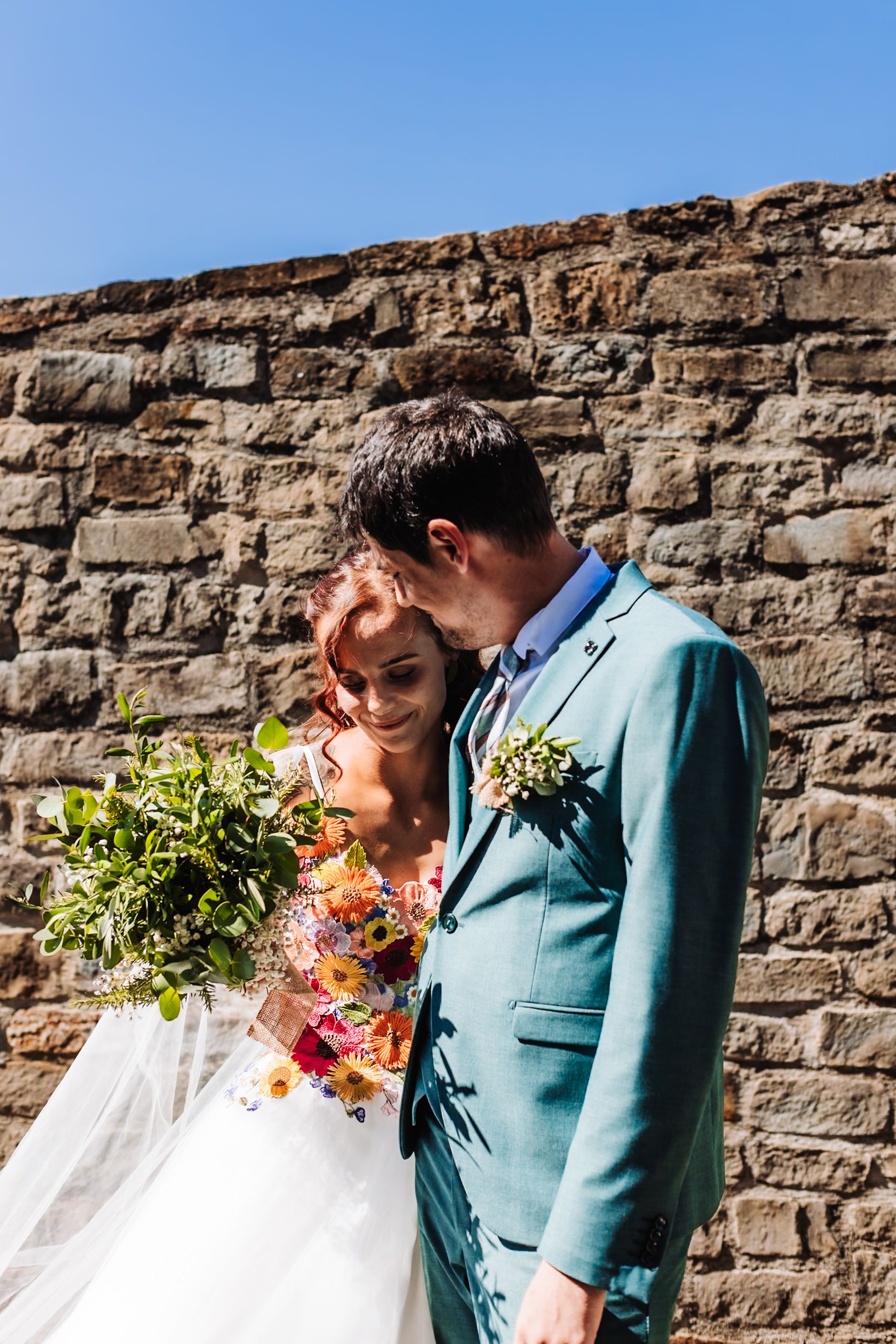 Bride and groom share a tender moment against a stone wall, she holds a colorful bouquet, he wears a teal suit.