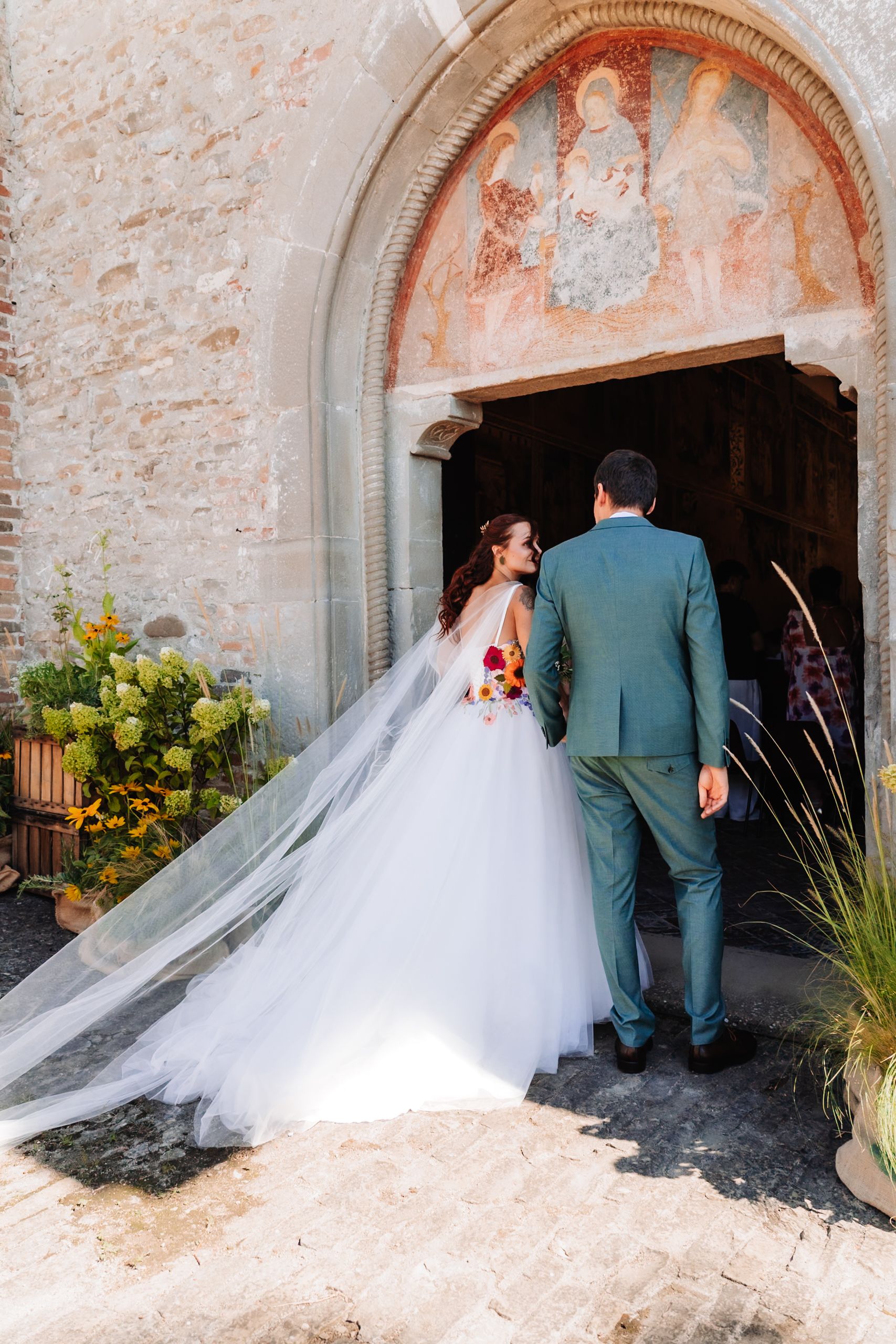 Bride and groom entering a historic stone church with fresco arch, bride in flowing white ballgown with long veil.
