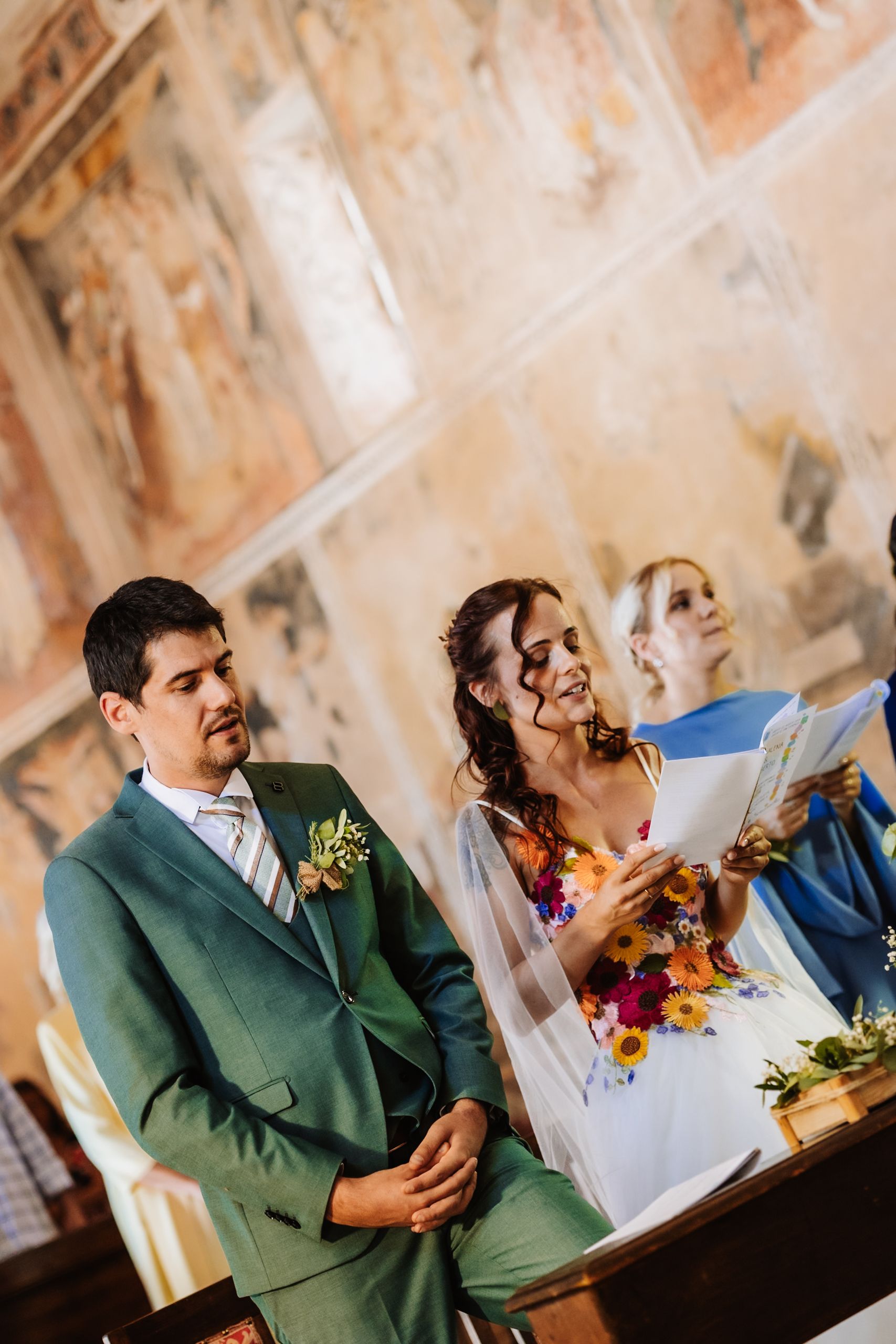 Bride smiling at groom during wedding ceremony in ornate chapel, with officiant holding papers in background.