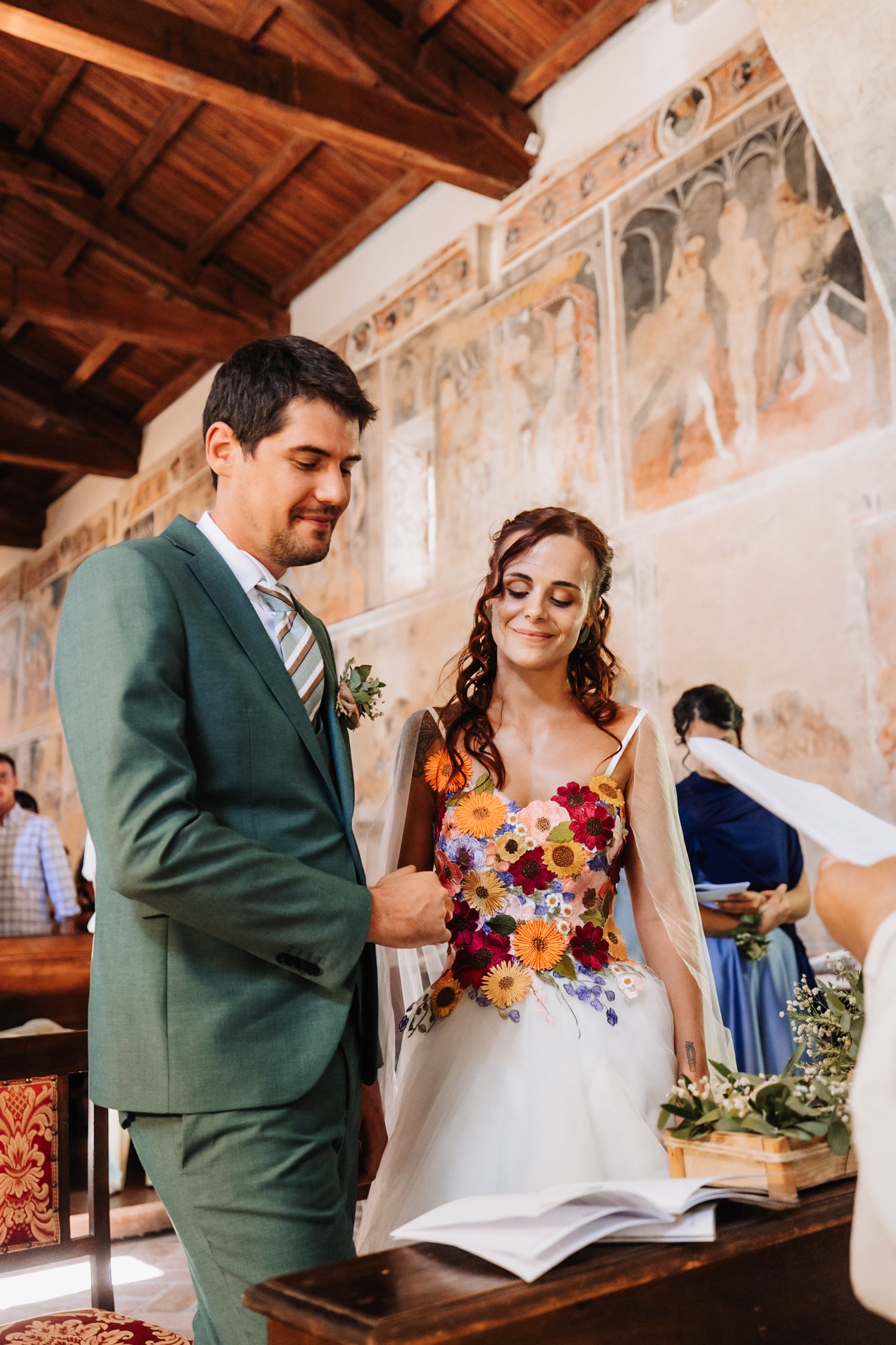 Bride in floral wedding dress and groom in green suit exchange vows in a historic church with fresco murals.