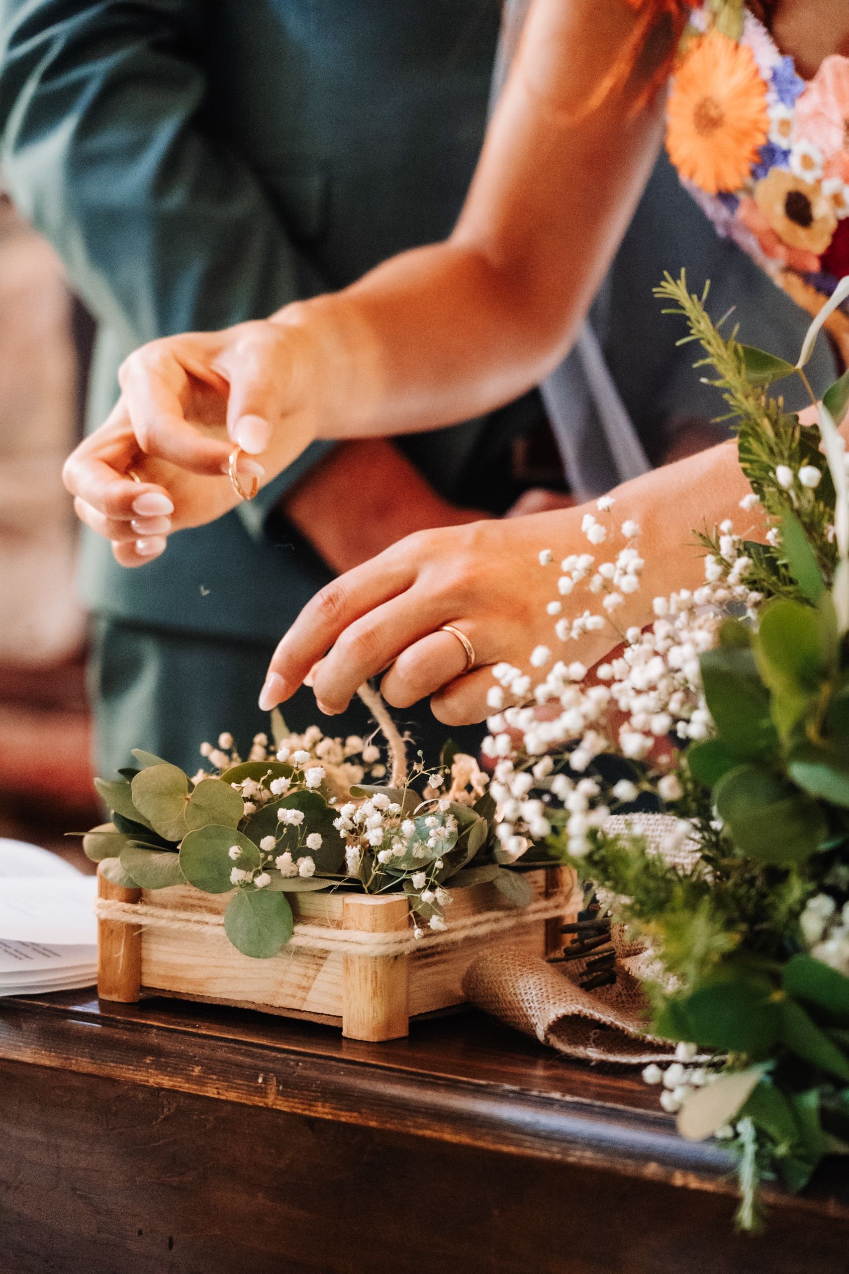 Two people arranging baby's breath and eucalyptus in a rustic wooden box centerpiece on a dark wood table.