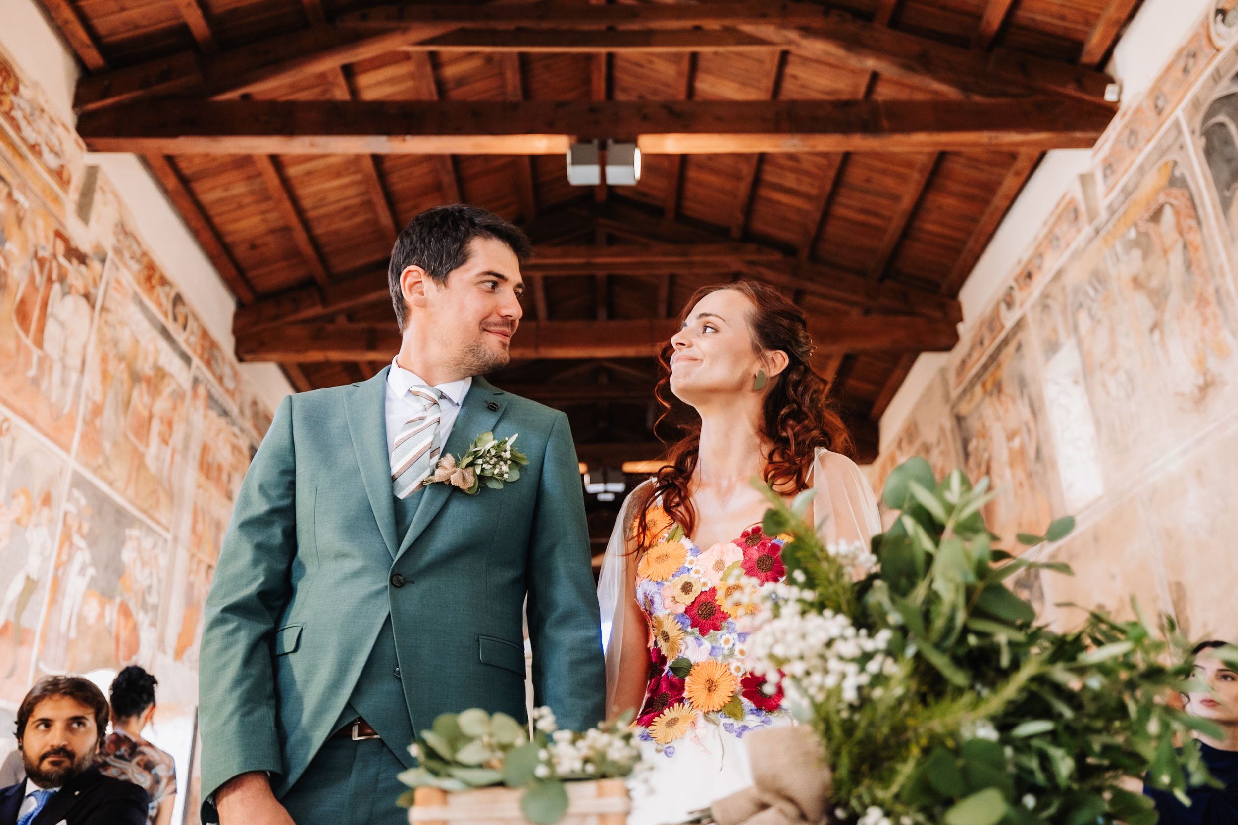 A couple shares a joyful moment at their wedding ceremony in a rustic venue with wooden ceiling and colorful frescoes.