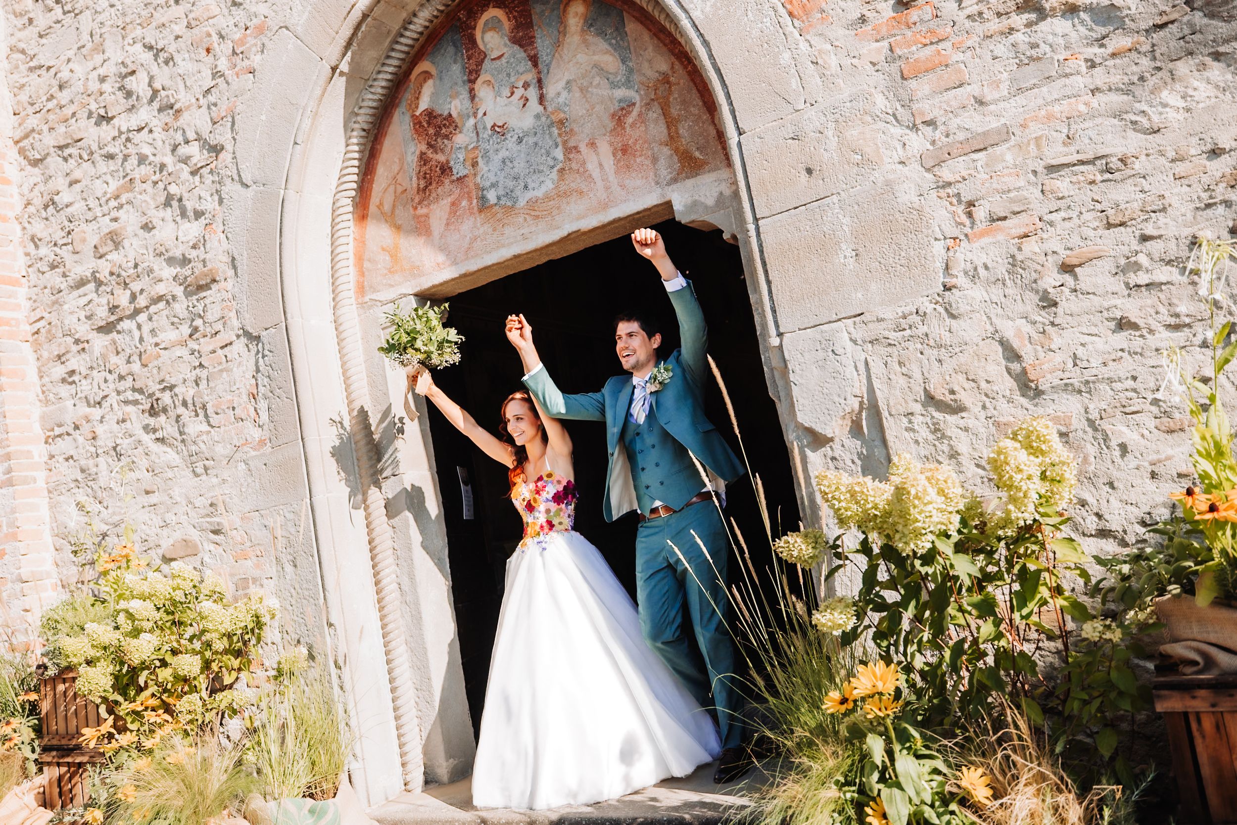 Joyful newlyweds celebrate at a stone church doorway, bride in white gown holding bouquet, groom in blue suit raising fists.