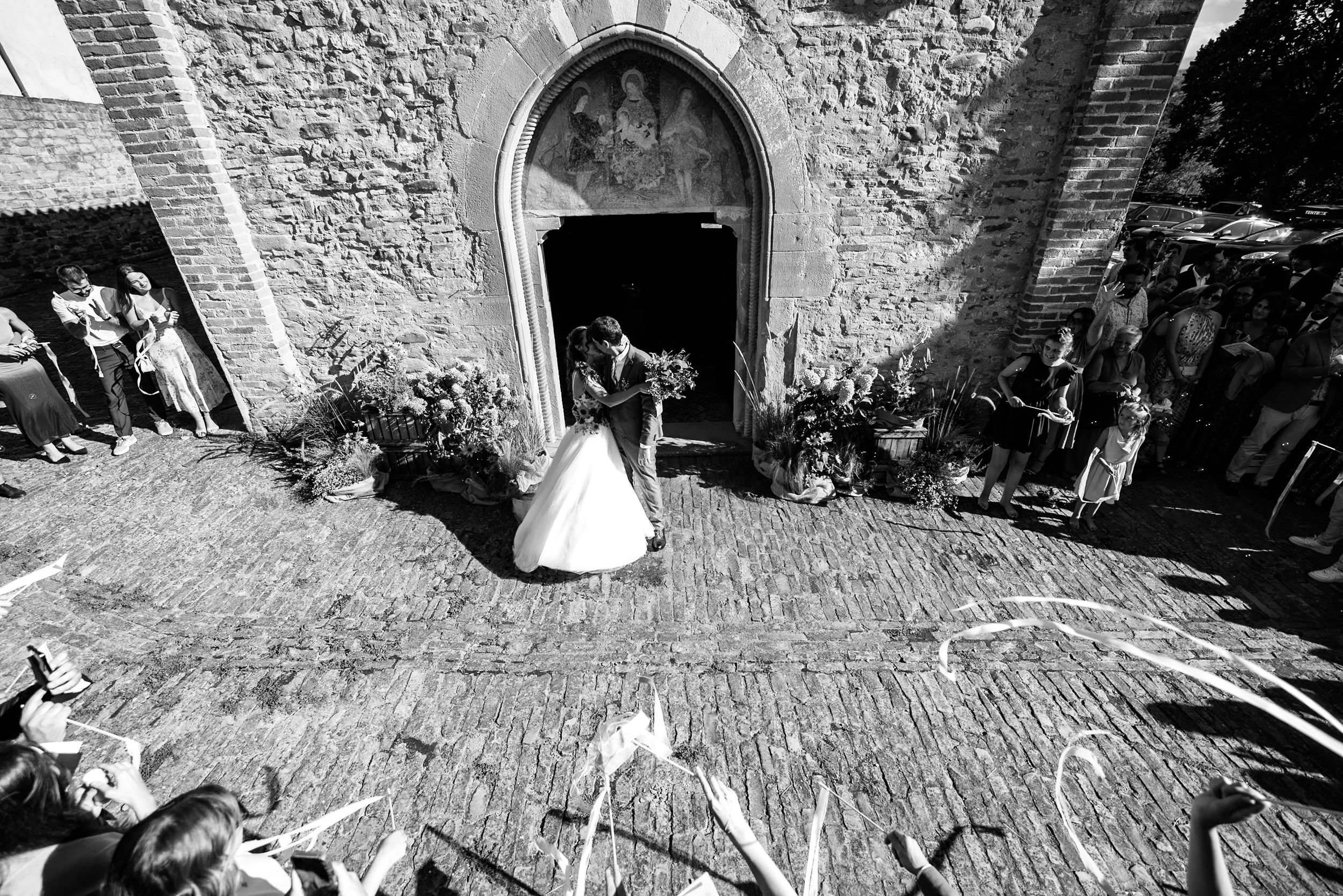Black and white photo of a bride and groom exiting a stone church as guests celebrate their wedding ceremony.