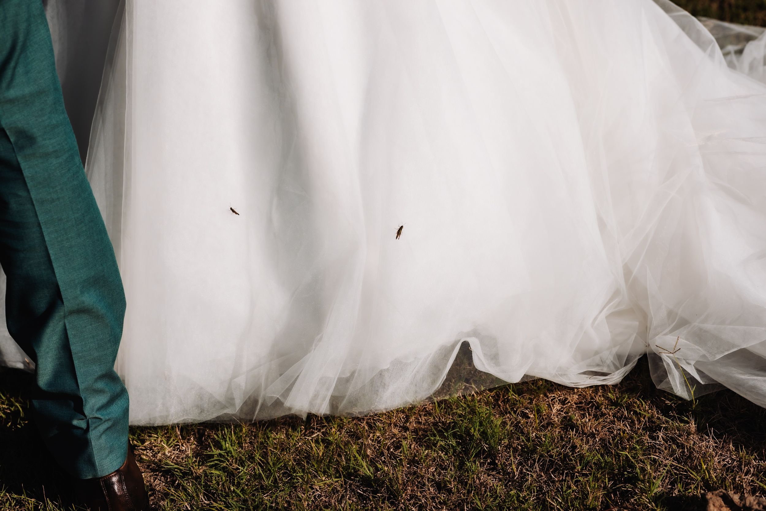 Close-up of a white tulle wedding dress hem with a groom in teal suit standing on green grass.