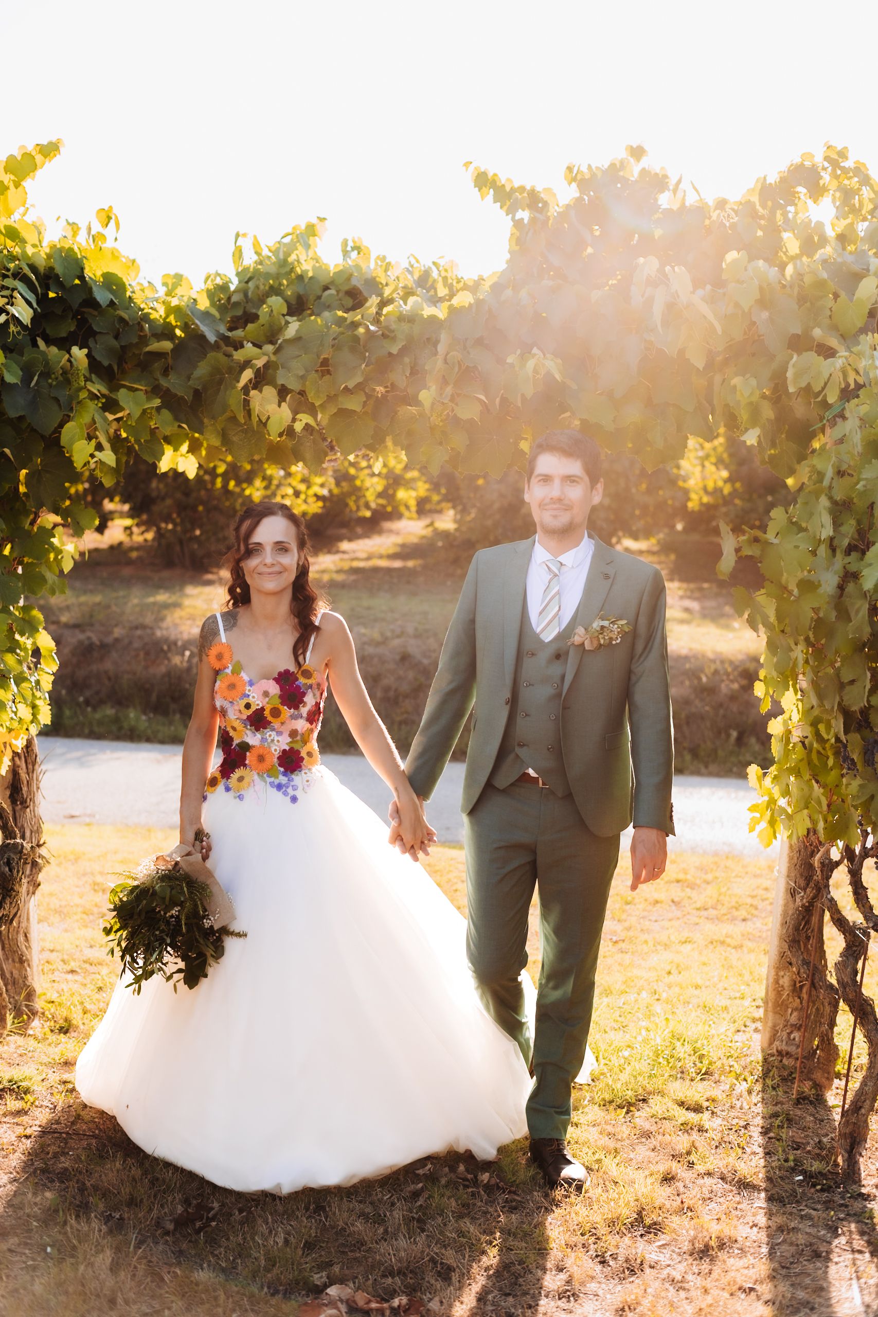 Bride in floral wedding dress holds groom's hand walking through vineyard at golden hour sunset.