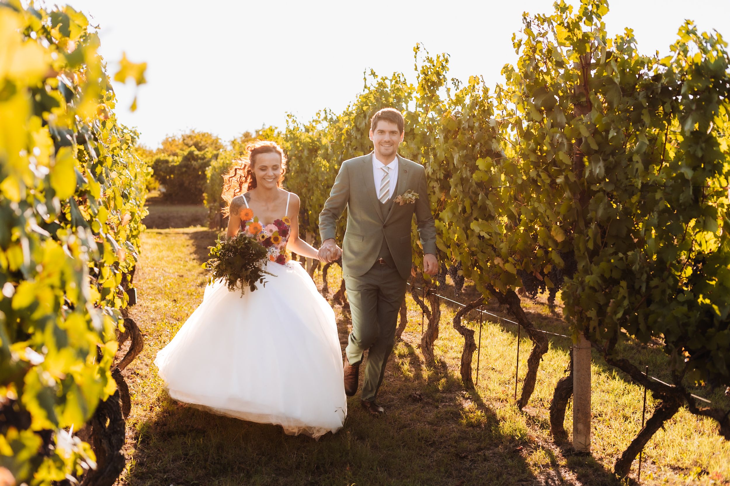 Bride and groom walking hand in hand through a sunlit vineyard, bride in white gown holding autumn bouquet.