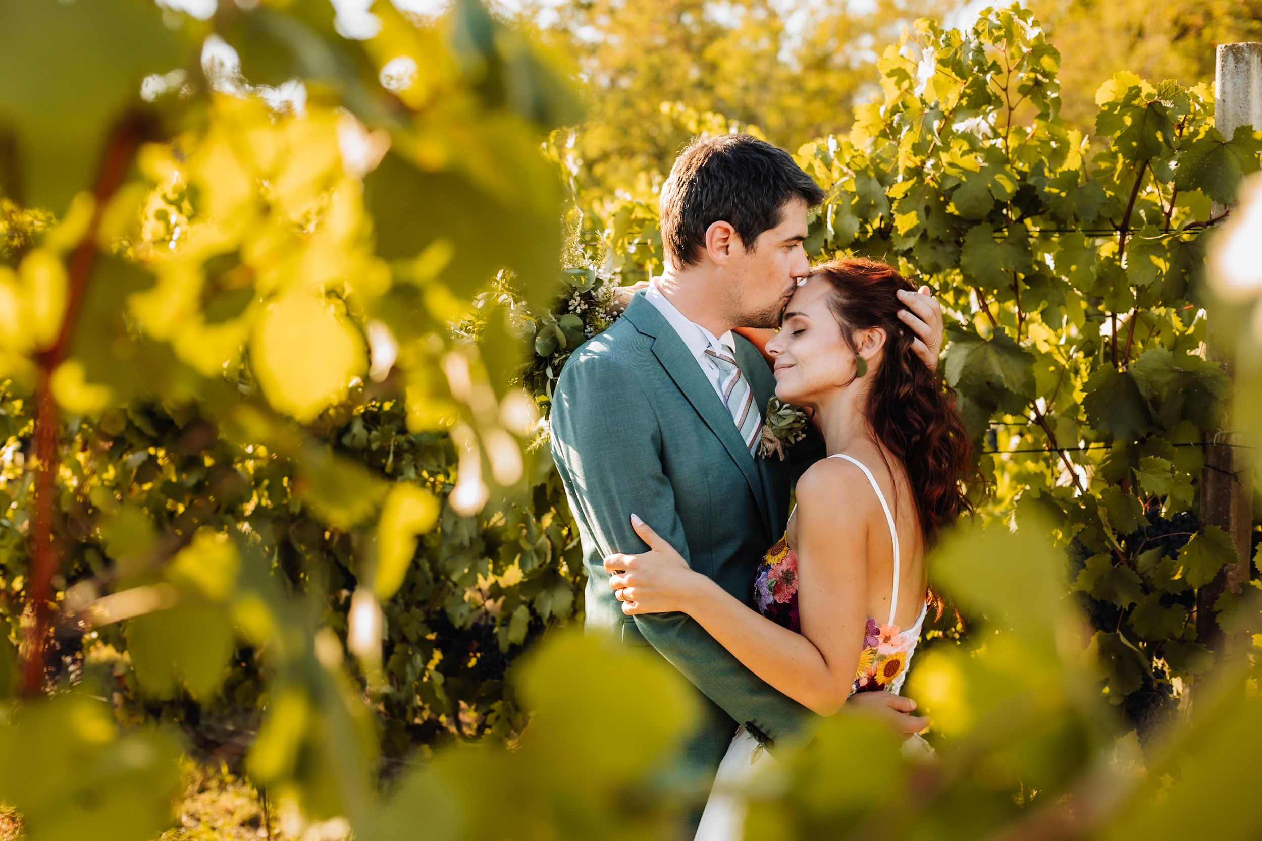 Couple embracing romantically among lush green grapevines at a vineyard wedding, man in teal suit, woman in floral dress.
