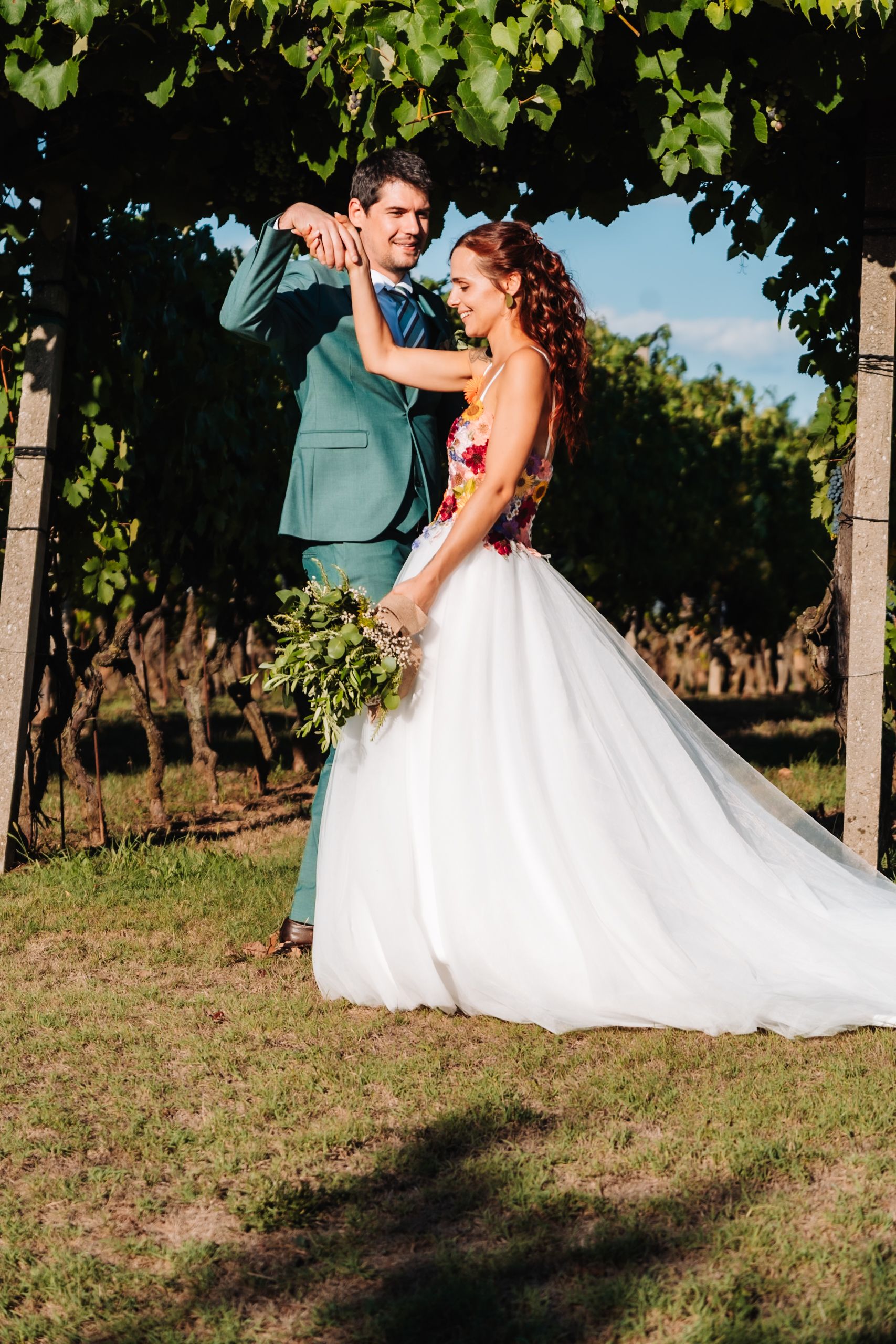 Bride in white floral gown and groom in teal suit dancing in a lush vineyard on their wedding day.