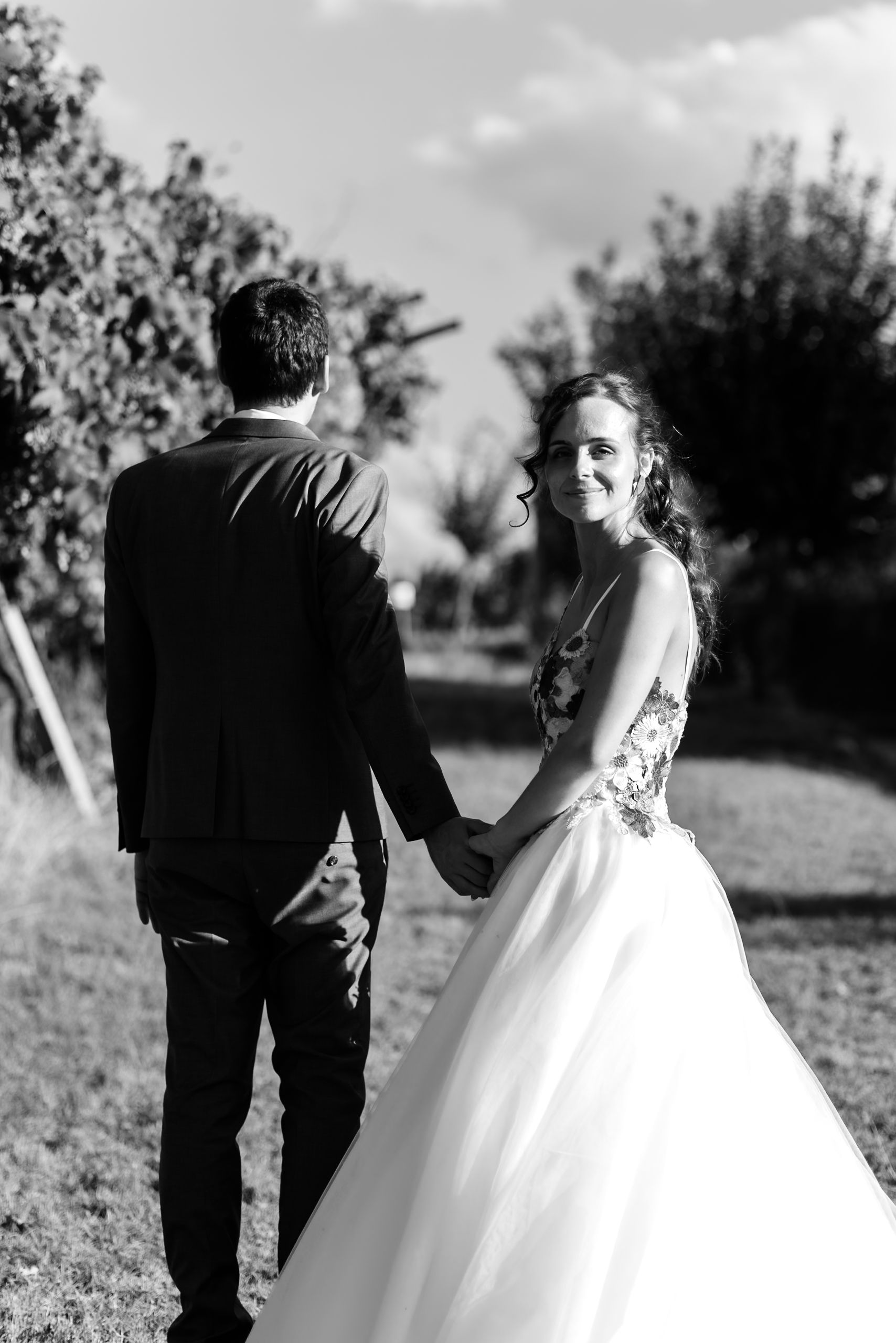 A bride in a floral gown holds hands with a groom in a suit while walking through a vineyard.