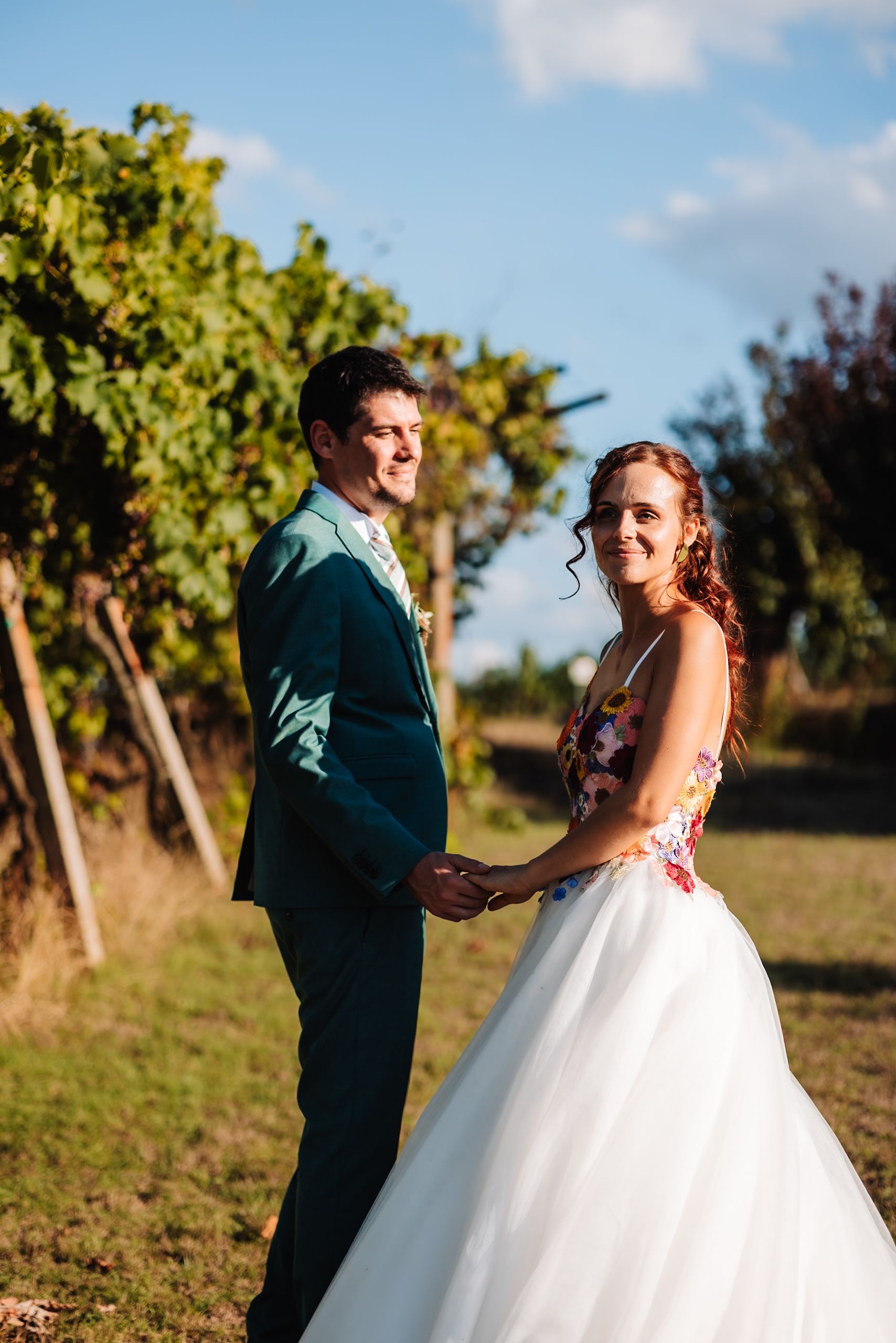 A smiling bride and groom hold hands in a sunlit vineyard, she in a floral gown and he in a teal suit.