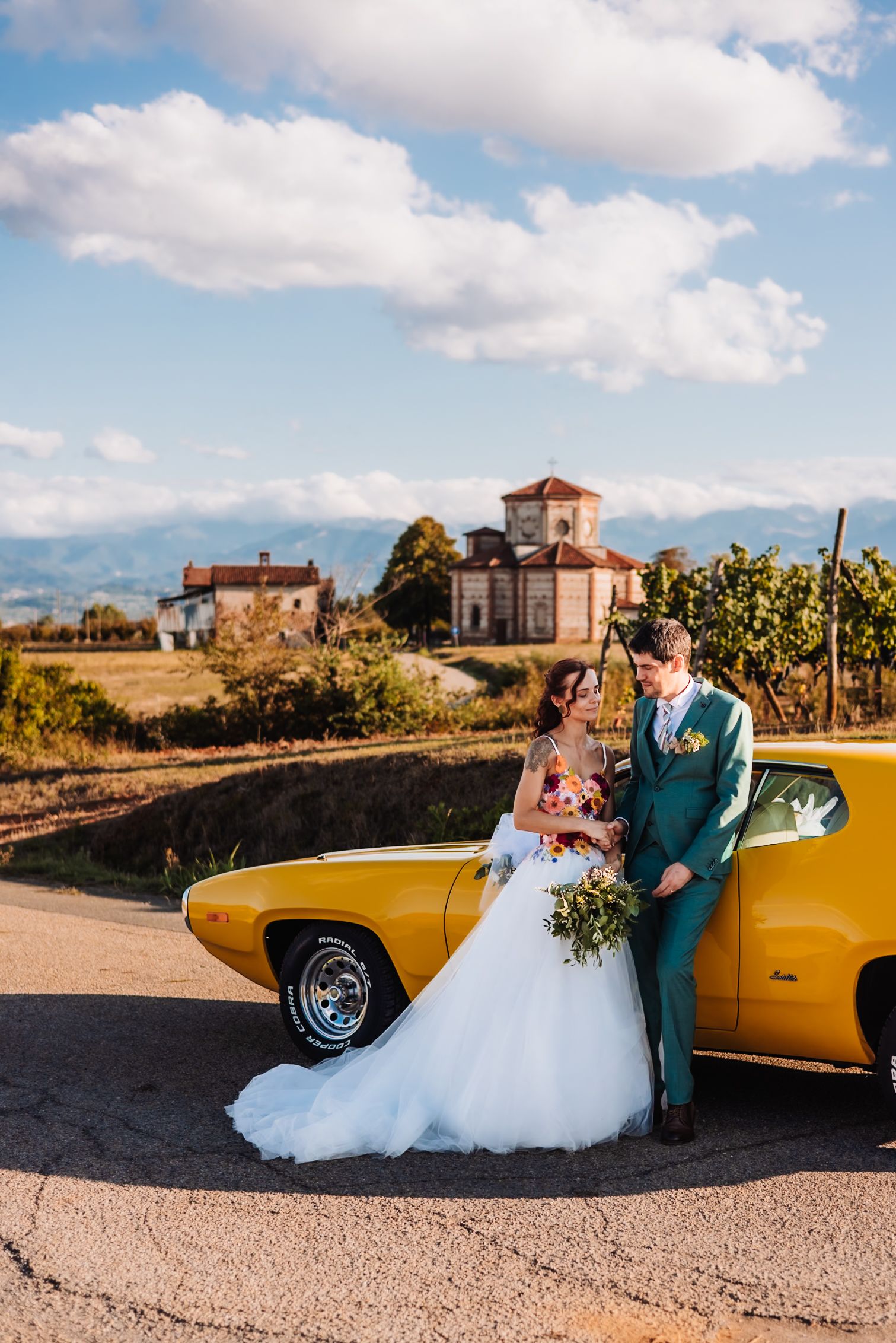 Bride and groom pose by a vintage yellow car at a vineyard estate on their wedding day.