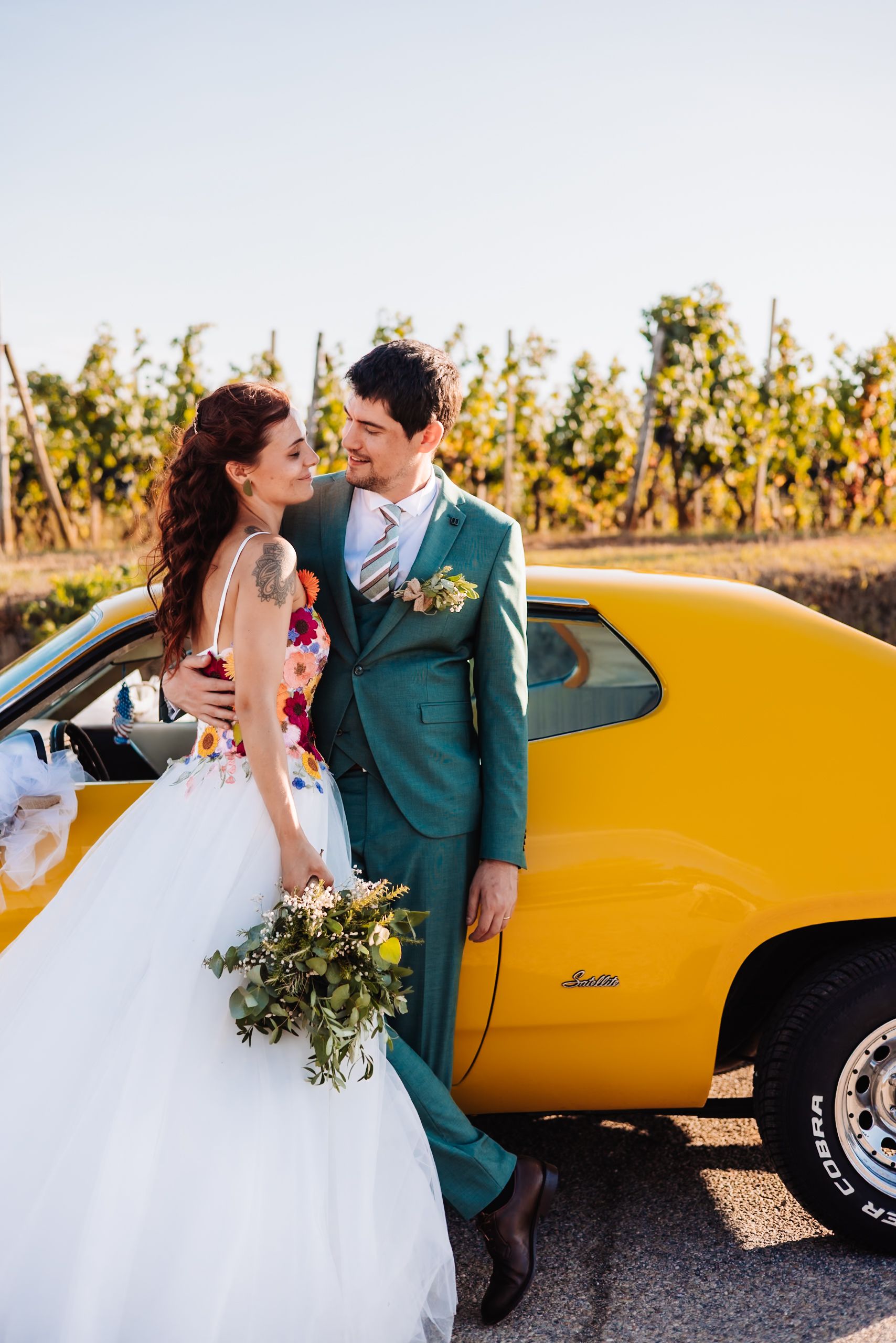 Bride and groom lean against a yellow classic car in a vineyard, wearing floral wedding attire and teal suit.