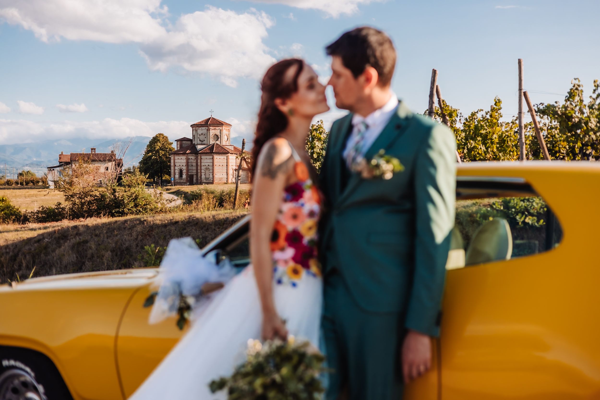 Couple kissing beside a vintage yellow car at a vineyard wedding with a rustic estate in the background.