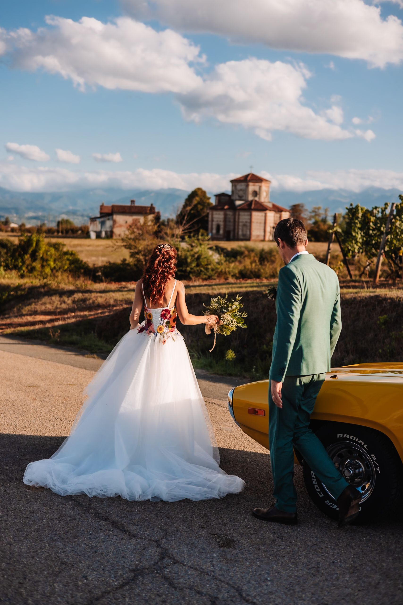 Bride in white gown and groom in teal suit walk toward a yellow classic car with a villa and mountains backdrop.