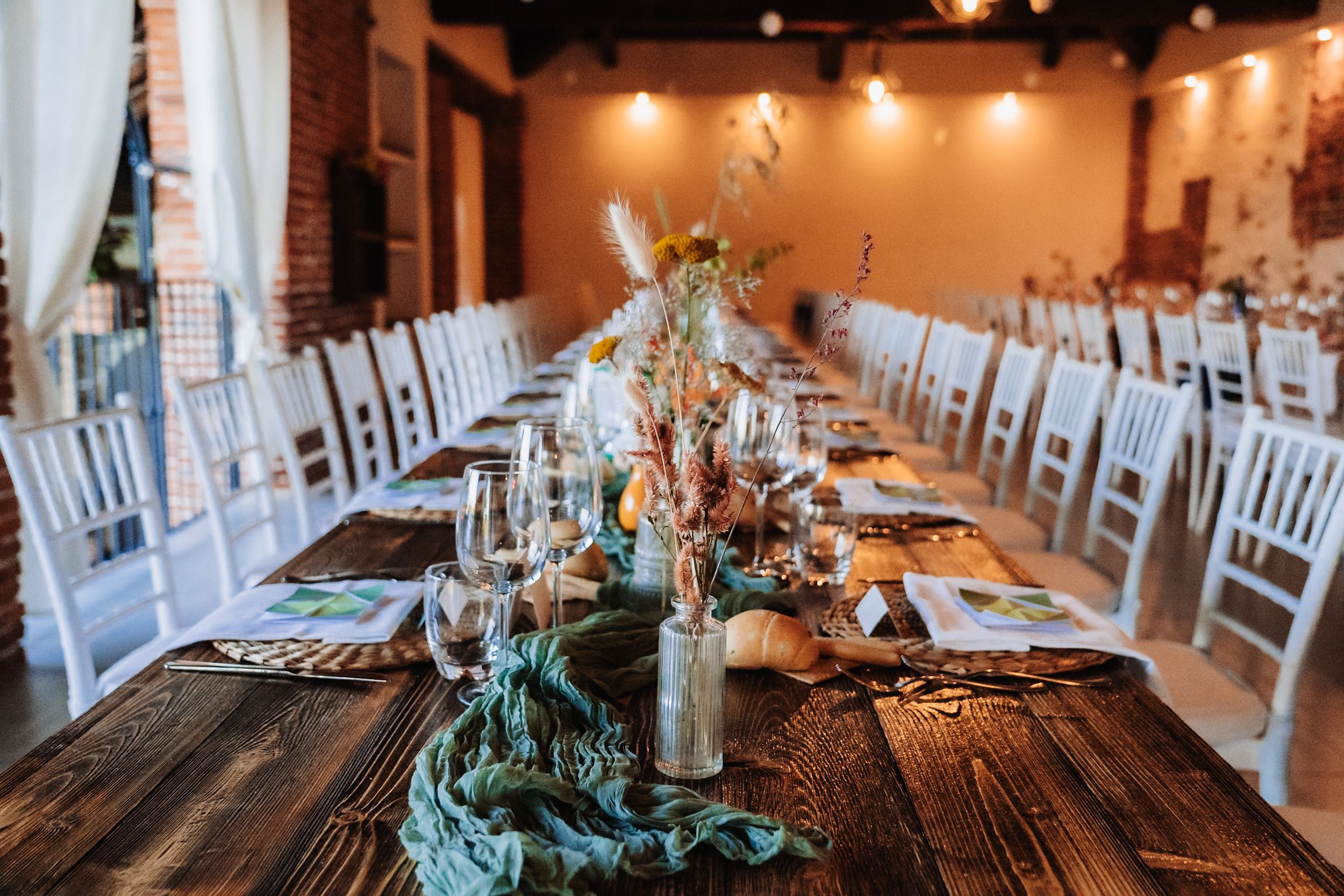 Rustic wooden banquet table with floral centerpieces, white chairs, and string lights in an elegant wedding reception venue.