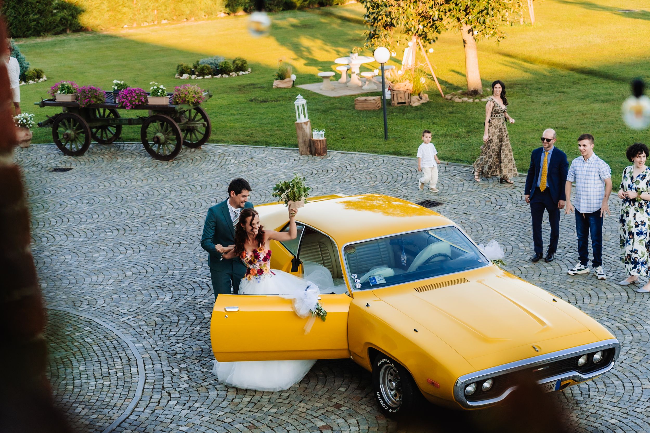 Bride and groom posing beside a vintage yellow muscle car at an elegant outdoor wedding venue.