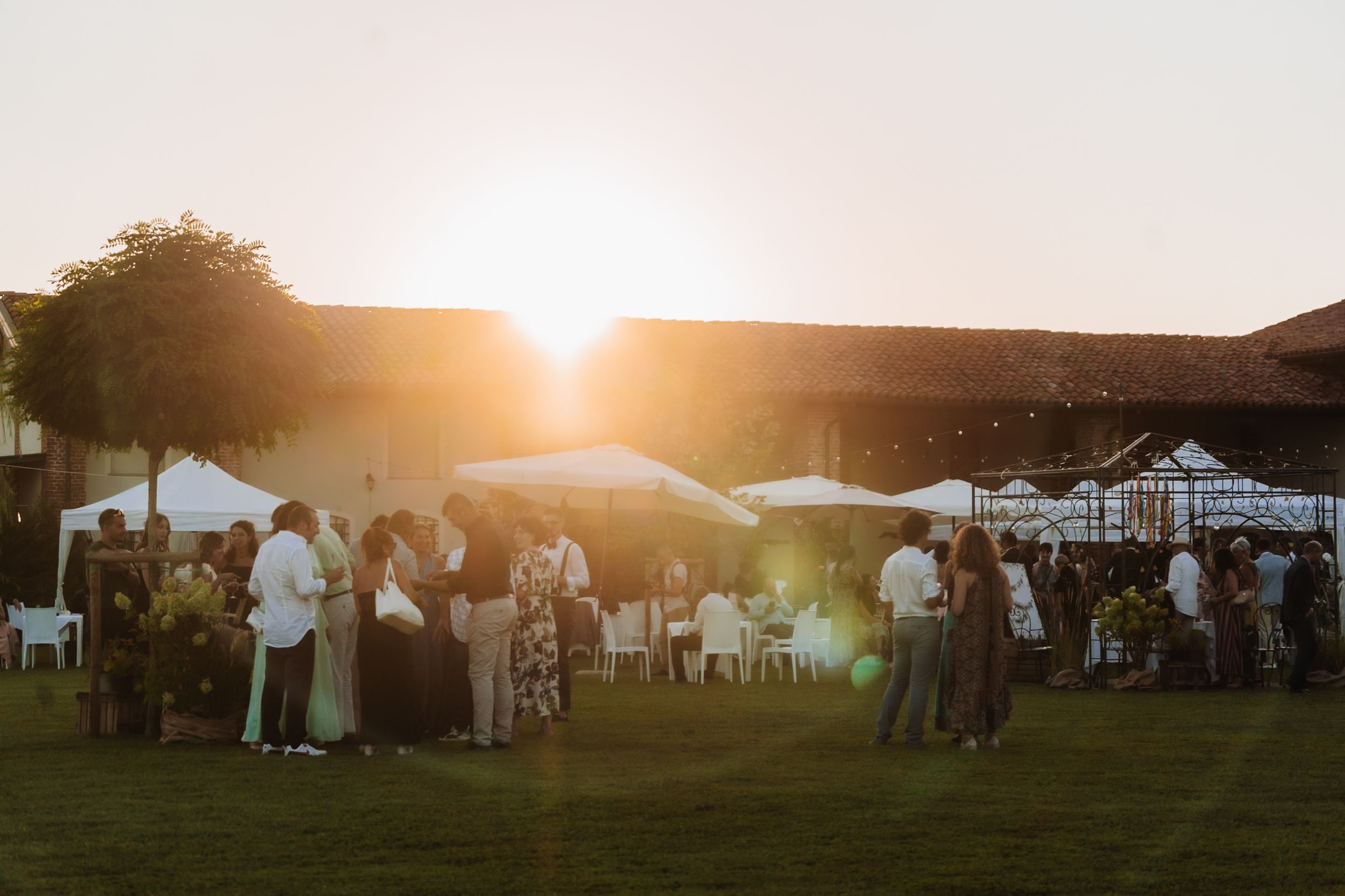 Guests mingle outdoors at a sunset garden party with white tents and warm golden light glowing behind a rustic barn.