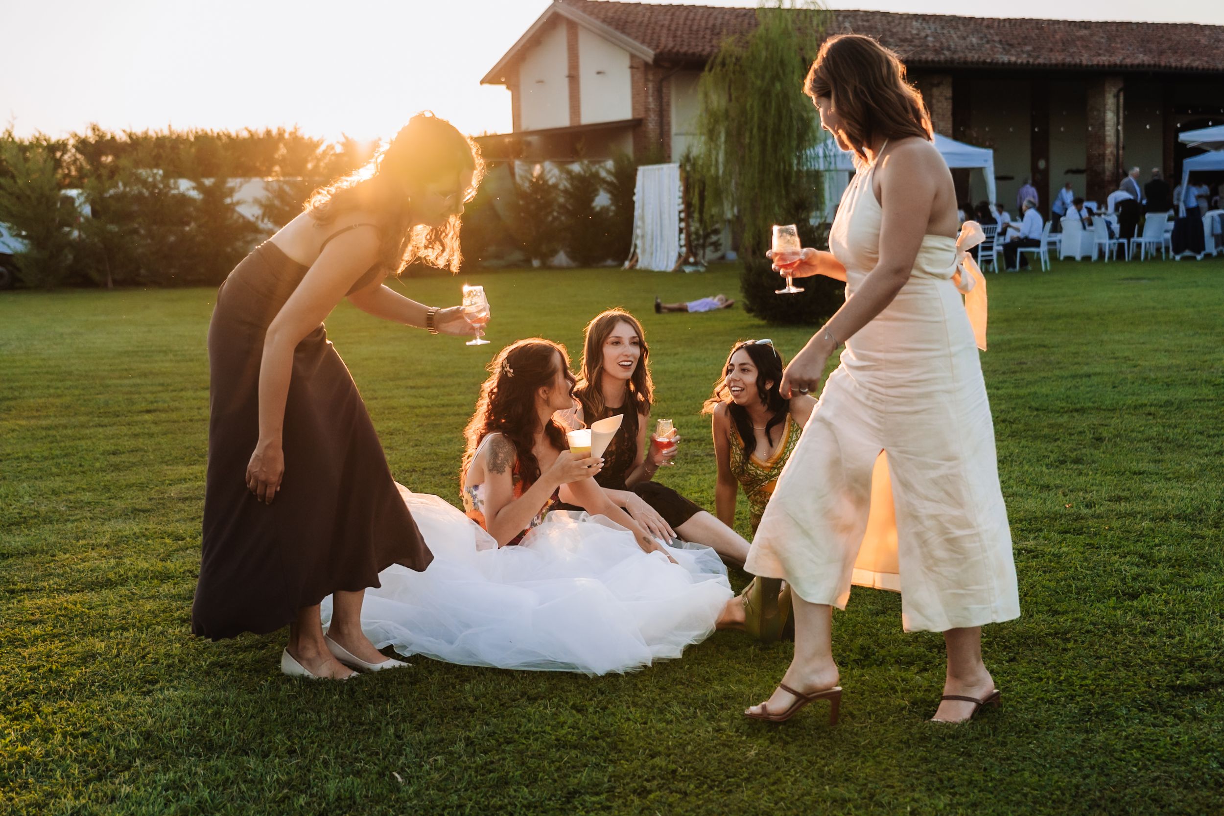 Bride in white gown celebrates with bridesmaids on green lawn at golden hour outdoor wedding reception.