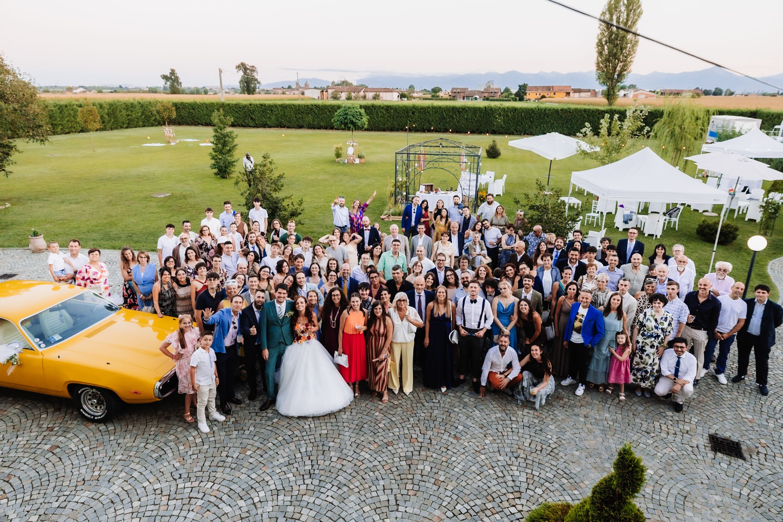 Large wedding group photo outdoors on cobblestone with lush green lawn, white tent, and vintage yellow car.