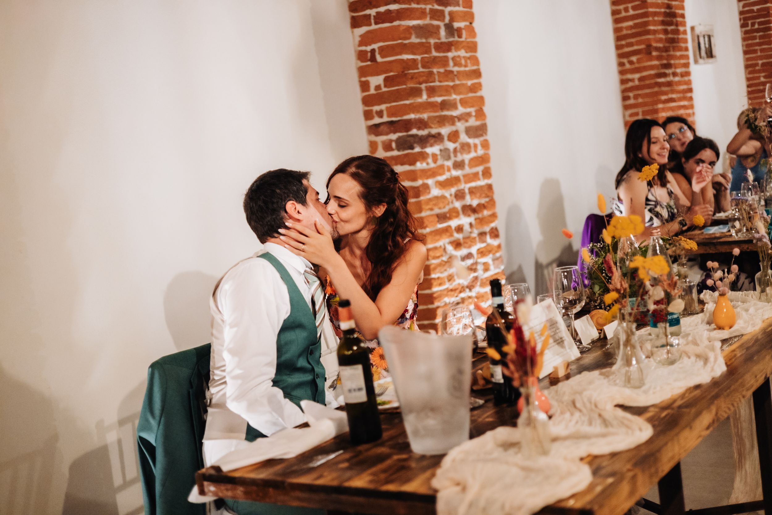 Bride and groom share a kiss at rustic wedding reception table with brick walls and floral centerpieces.