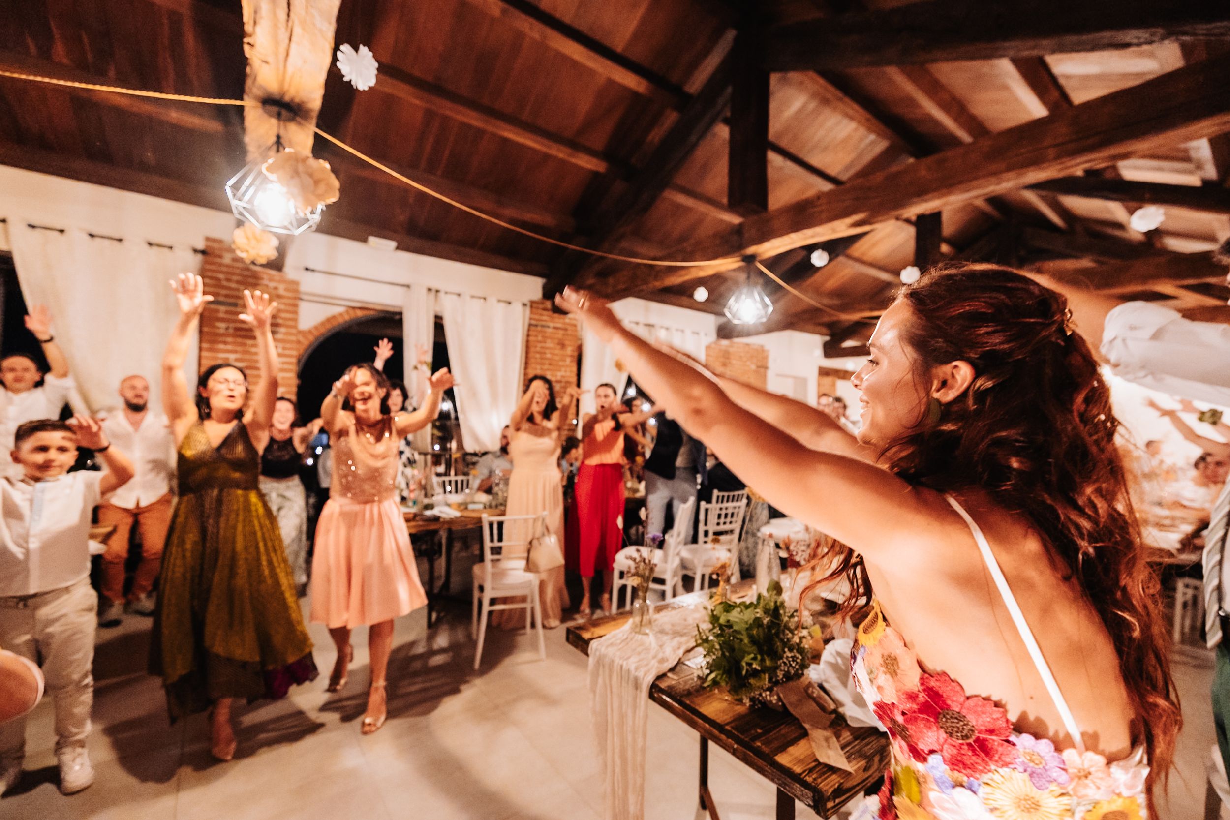 Bride tosses bouquet to excited wedding guests in a decorated reception venue with string lights.