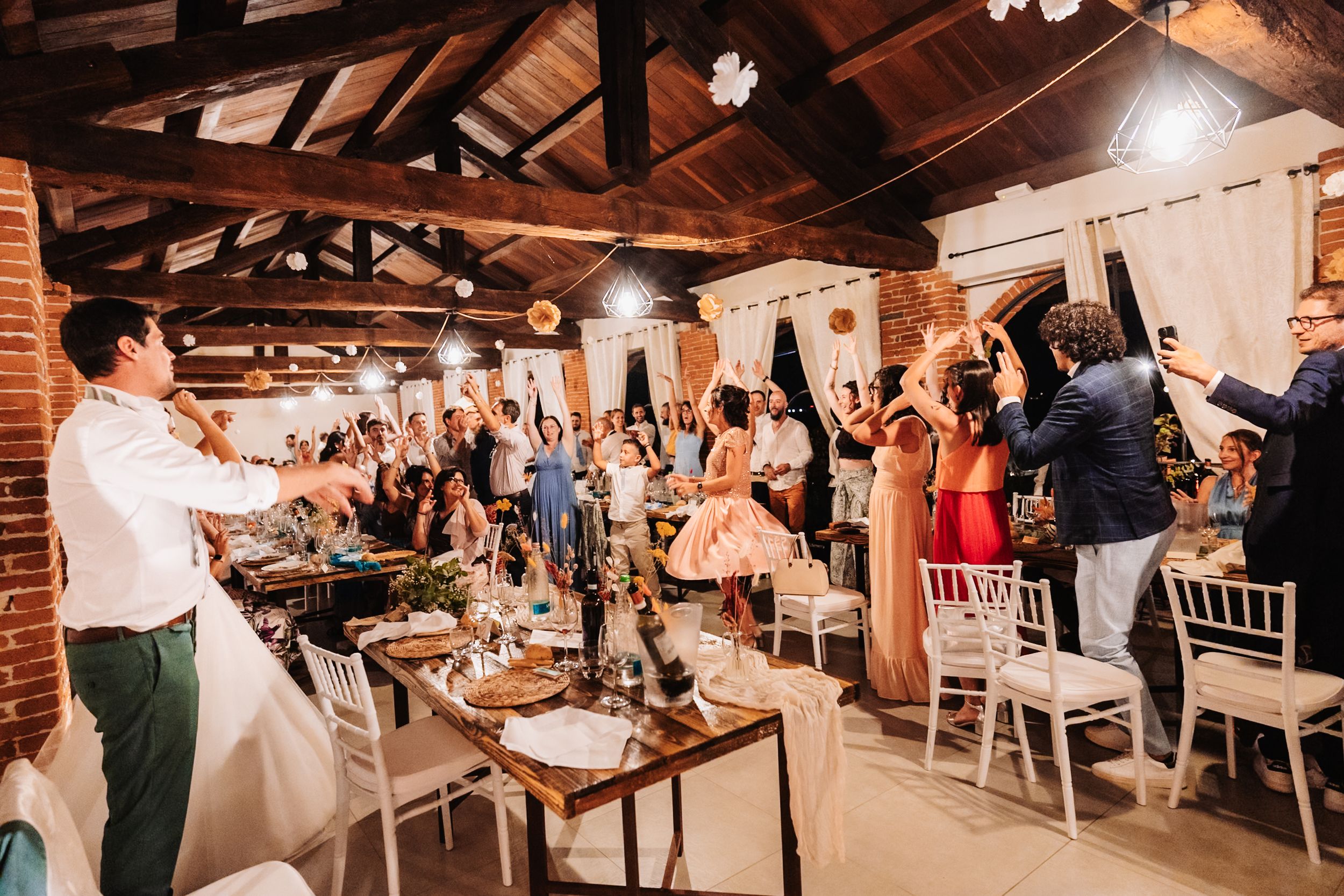 Wedding reception guests raising glasses in a rustic venue with wooden beams, white chairs, and warm festive lighting.
