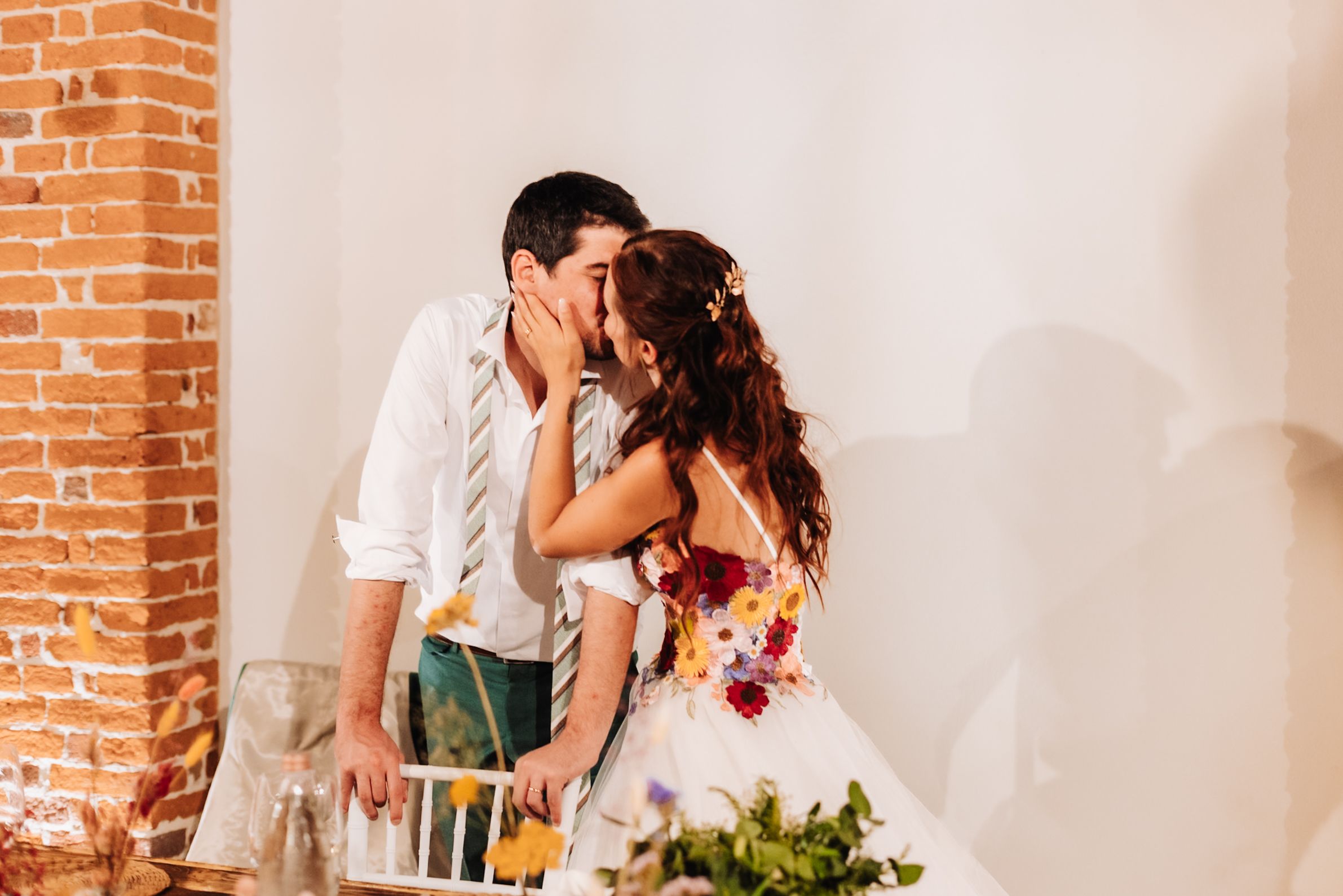 Bride and groom share a romantic kiss at their wedding reception near a rustic brick wall.
