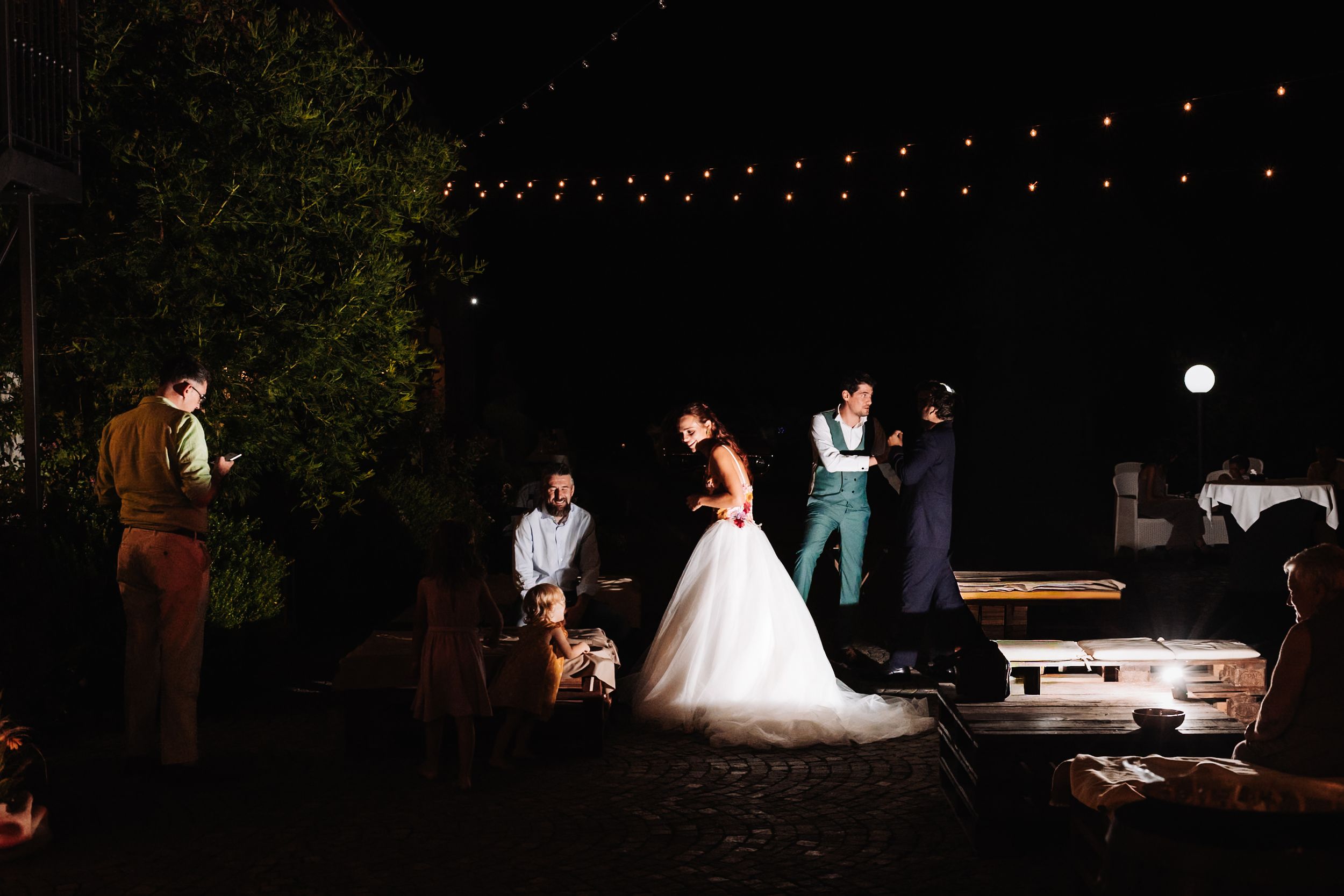 Bride and groom share a dance outdoors at night, surrounded by guests under warm string lights.