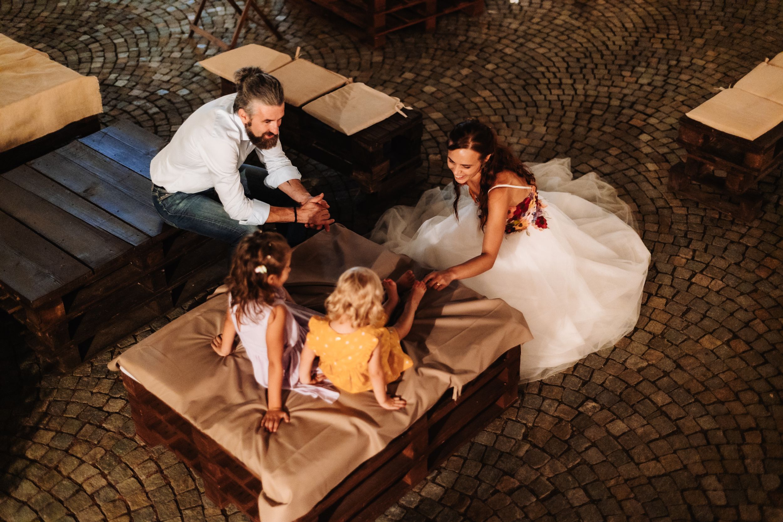 Bride and groom sitting with two young girls on wooden furniture at an outdoor wedding reception.