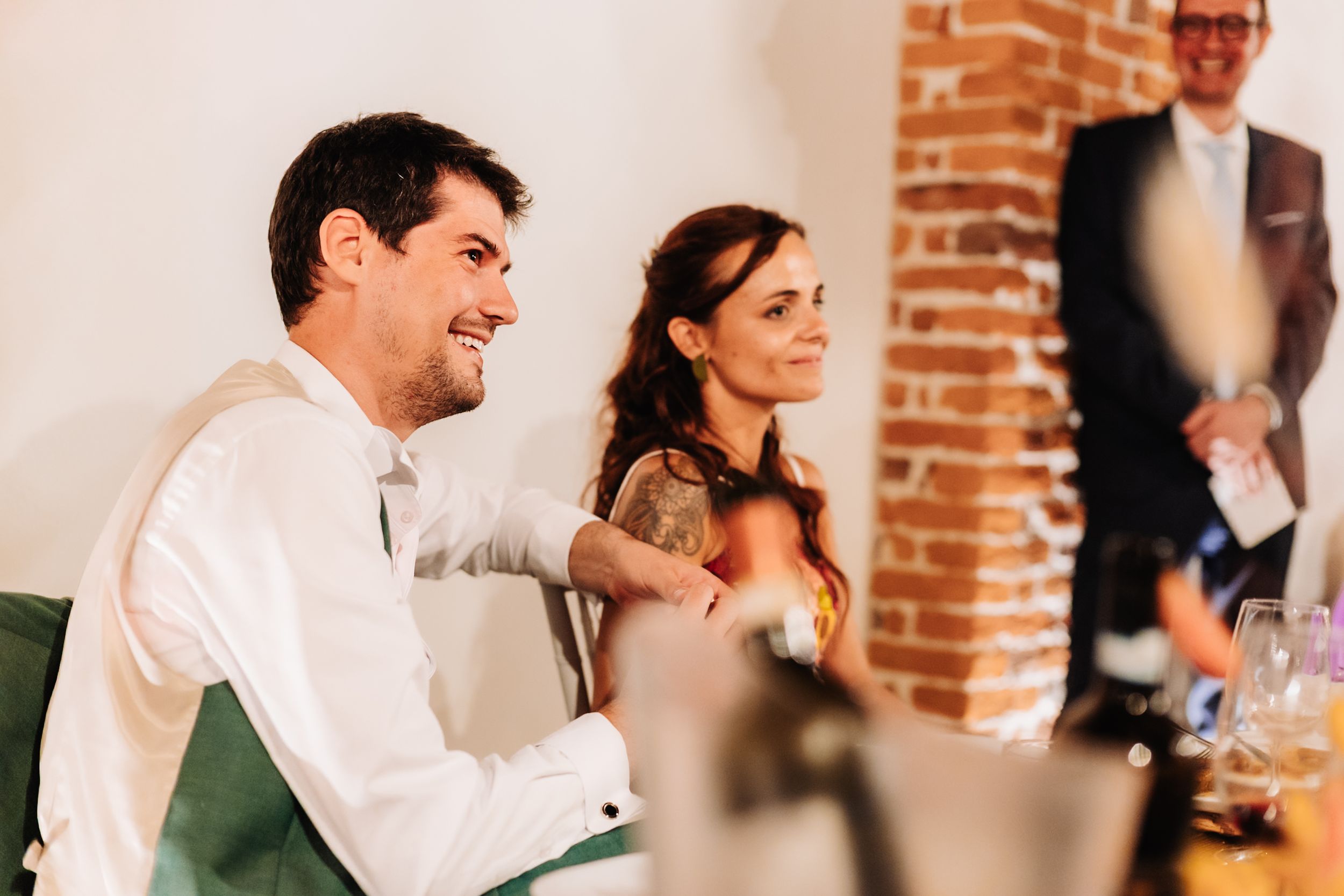 Smiling couple seated at a wedding reception, man in white shirt laughing, brick wall background.