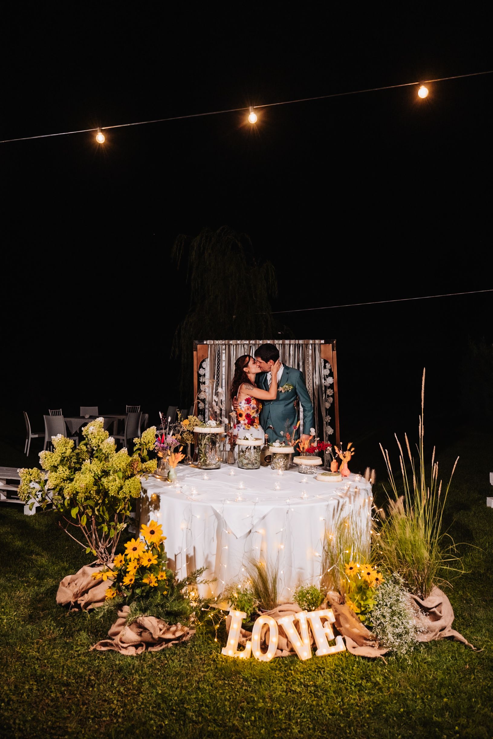 Couple kissing at outdoor wedding sweetheart table with sunflowers, string lights, and illuminated LOVE sign at night.