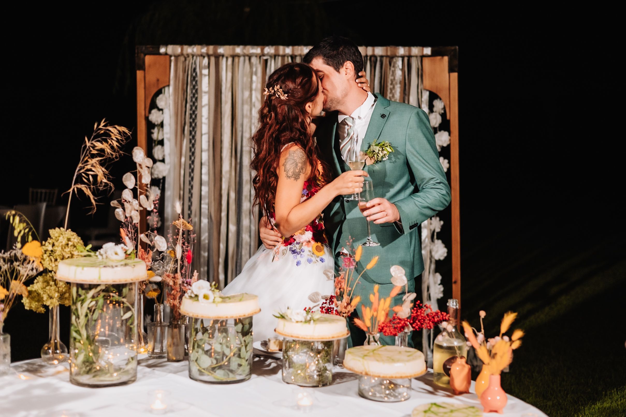 Couple kissing at wedding reception with floral arrangements and terrariums on display table at night.