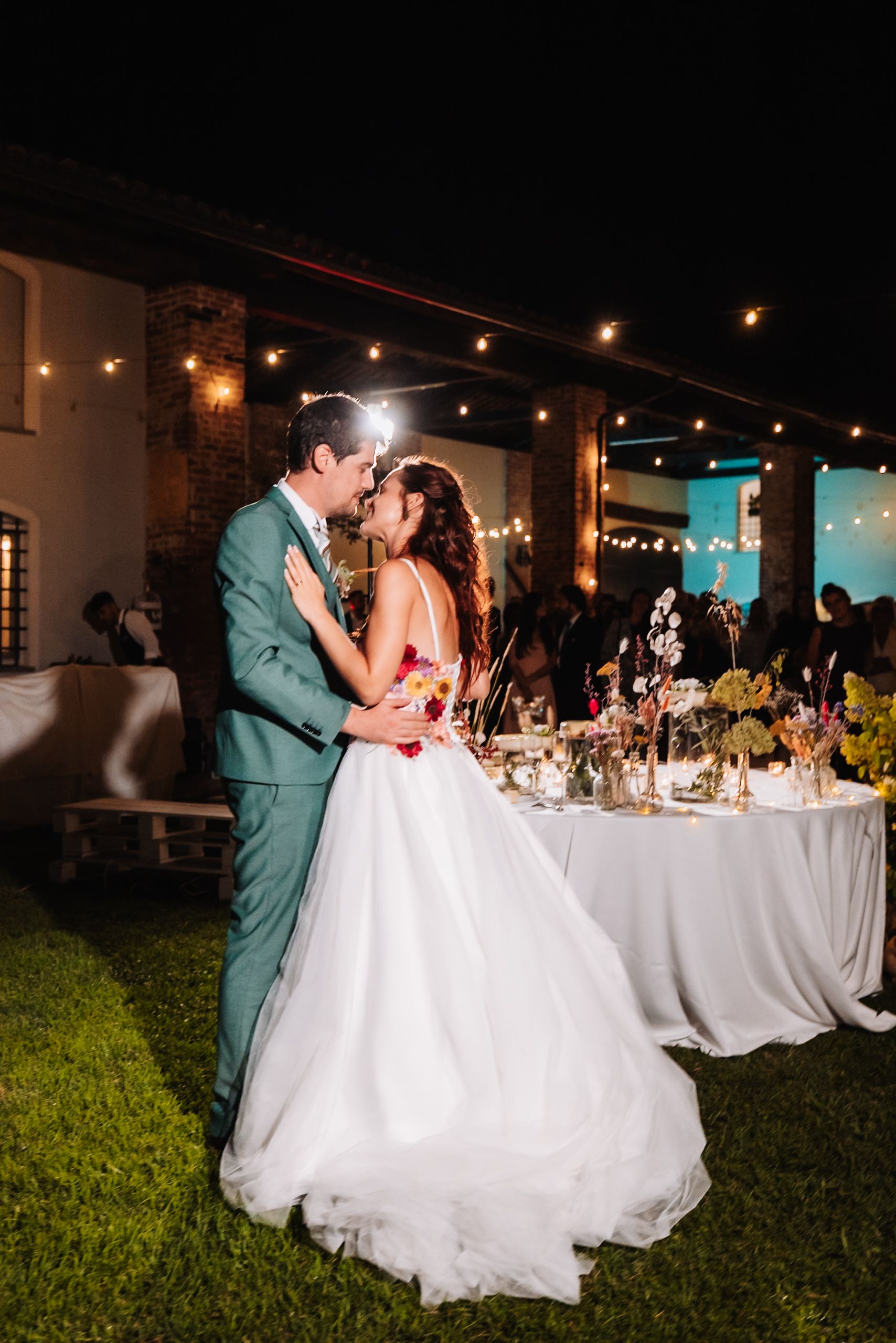 Bride and groom share first dance outdoors at night under string lights at elegant wedding reception.
