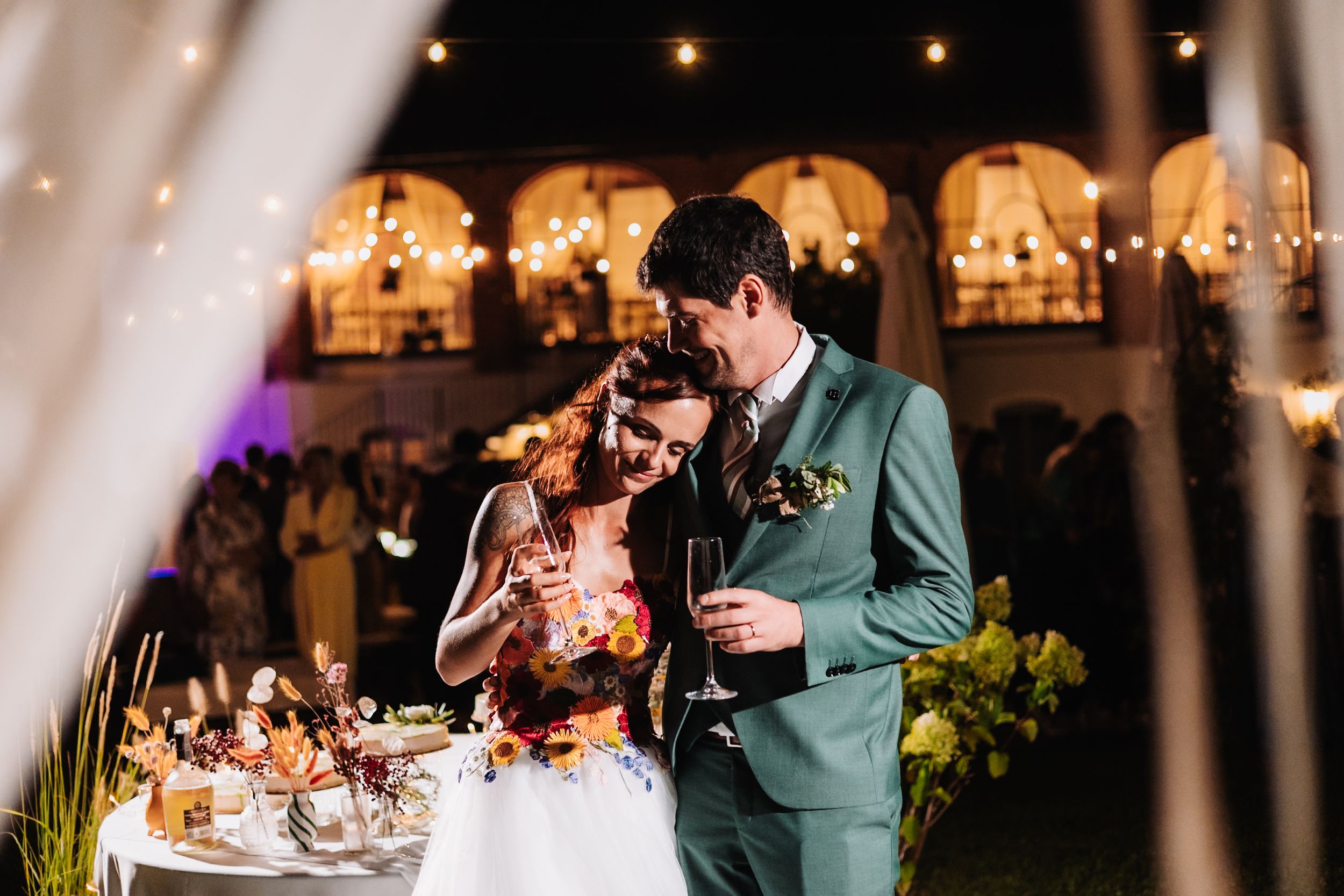 Bride and groom share a romantic toast at an outdoor evening wedding reception with warm string lights.