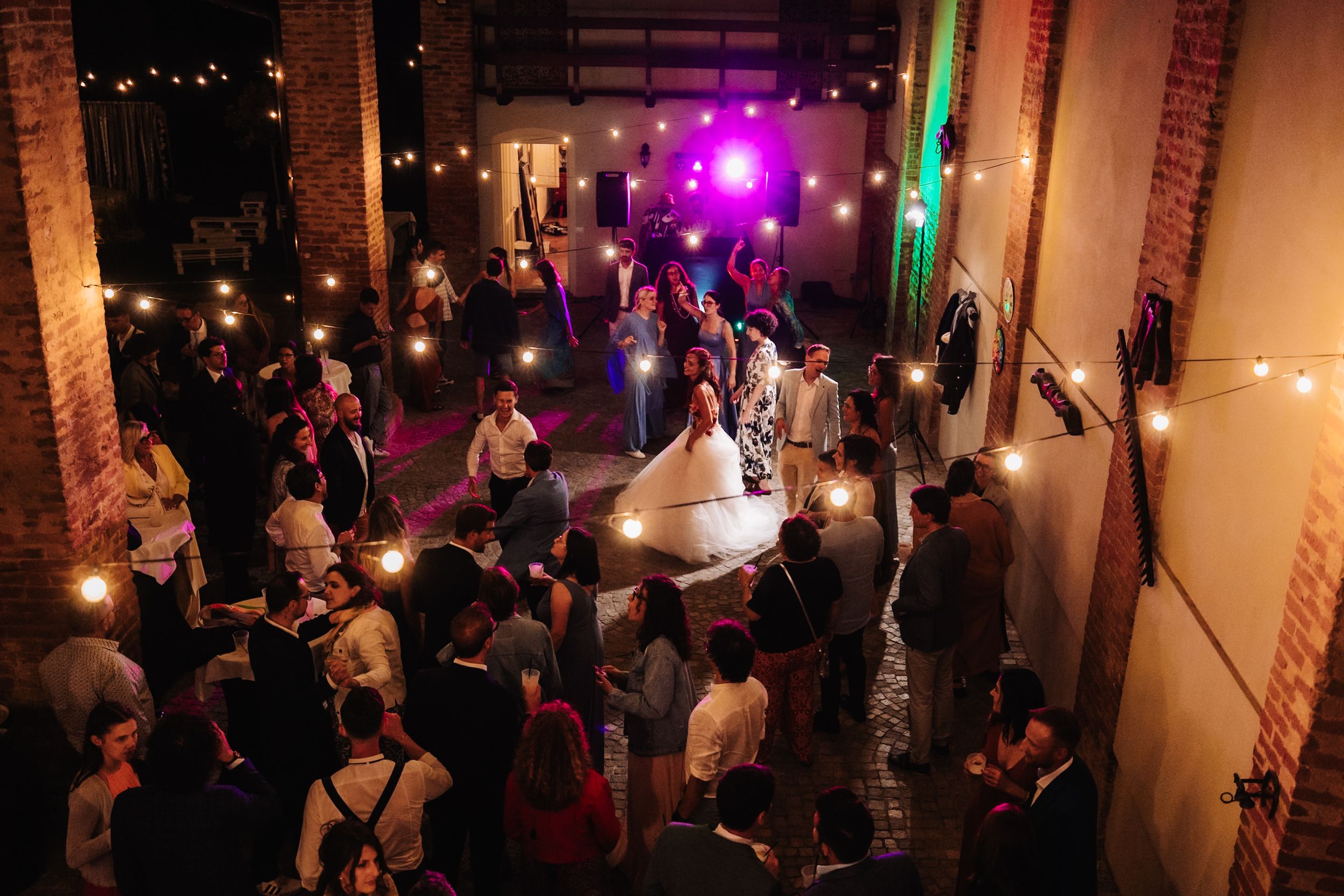 Aerial view of wedding reception with bride and groom dancing surrounded by guests under warm string lights in rustic venue.