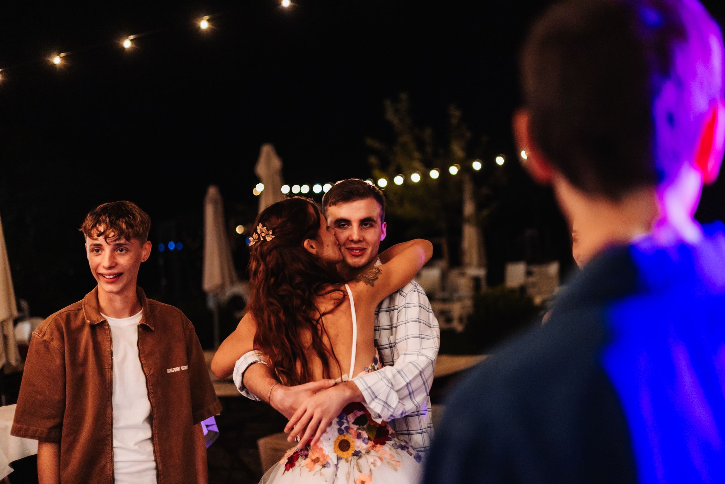 Young people hugging and dancing outdoors at a night party with string lights in the background.