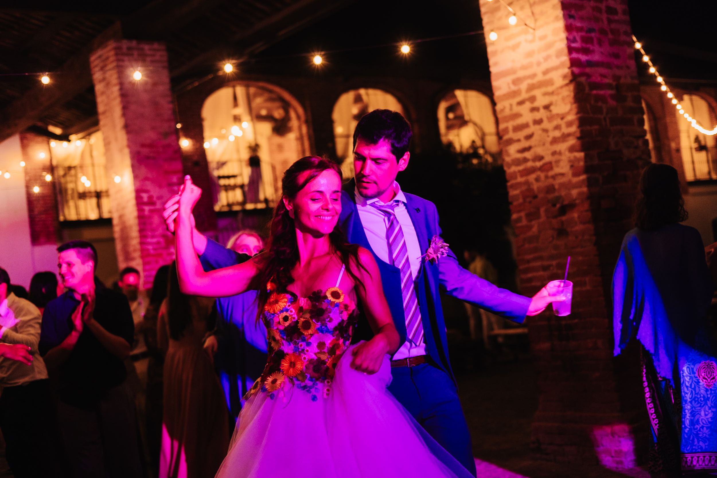 Bride and groom share a joyful first dance at their wedding reception under warm string lights in a rustic brick venue.