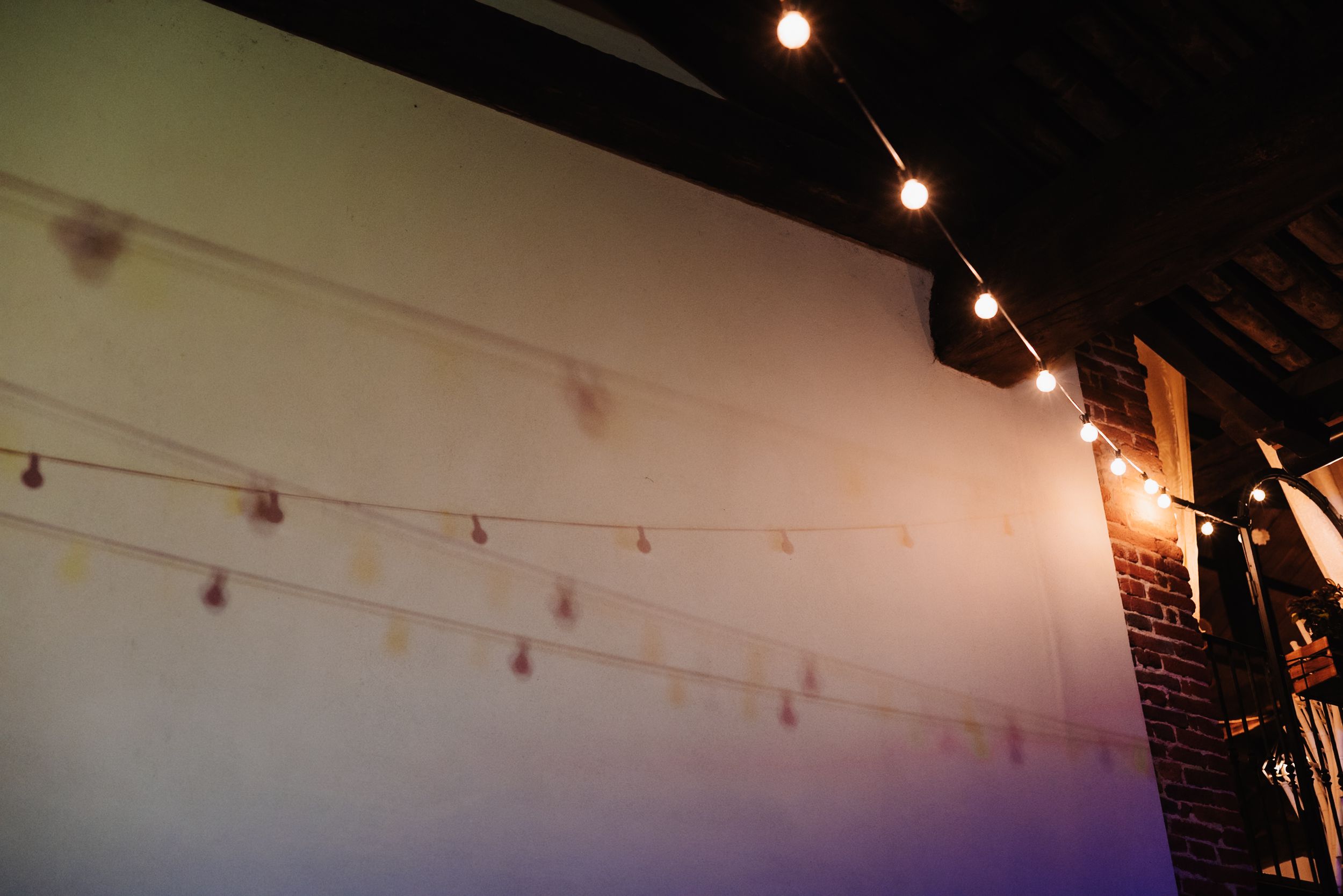 String lights draped across a white wall in a dimly lit venue with brick accents and track lighting above.