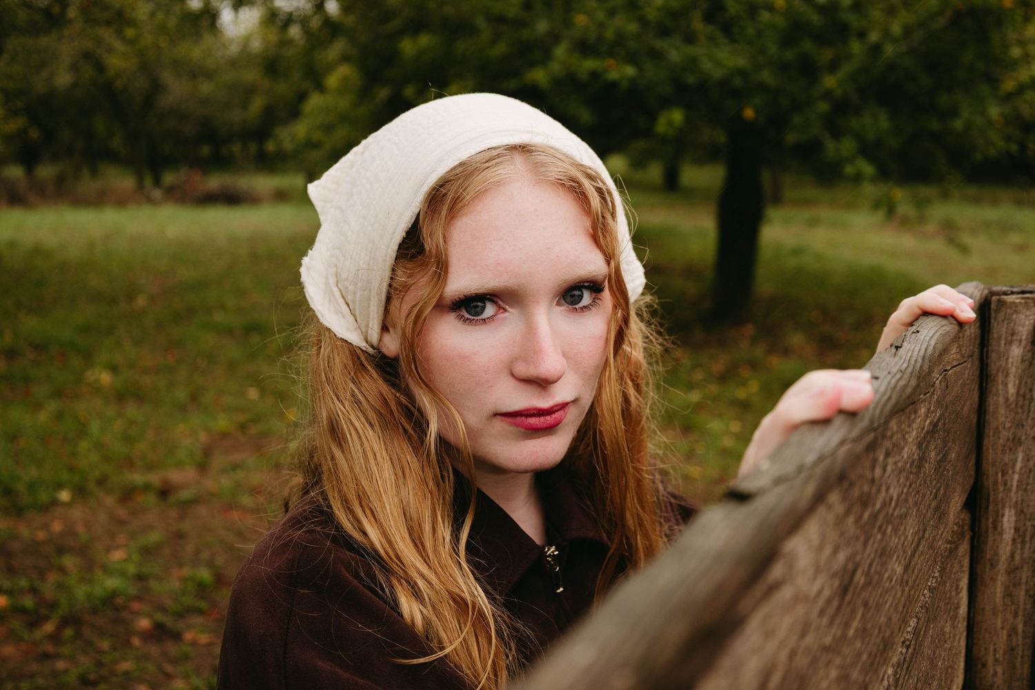 Young woman with red hair and white headscarf leans on wooden fence in a lush green orchard setting.