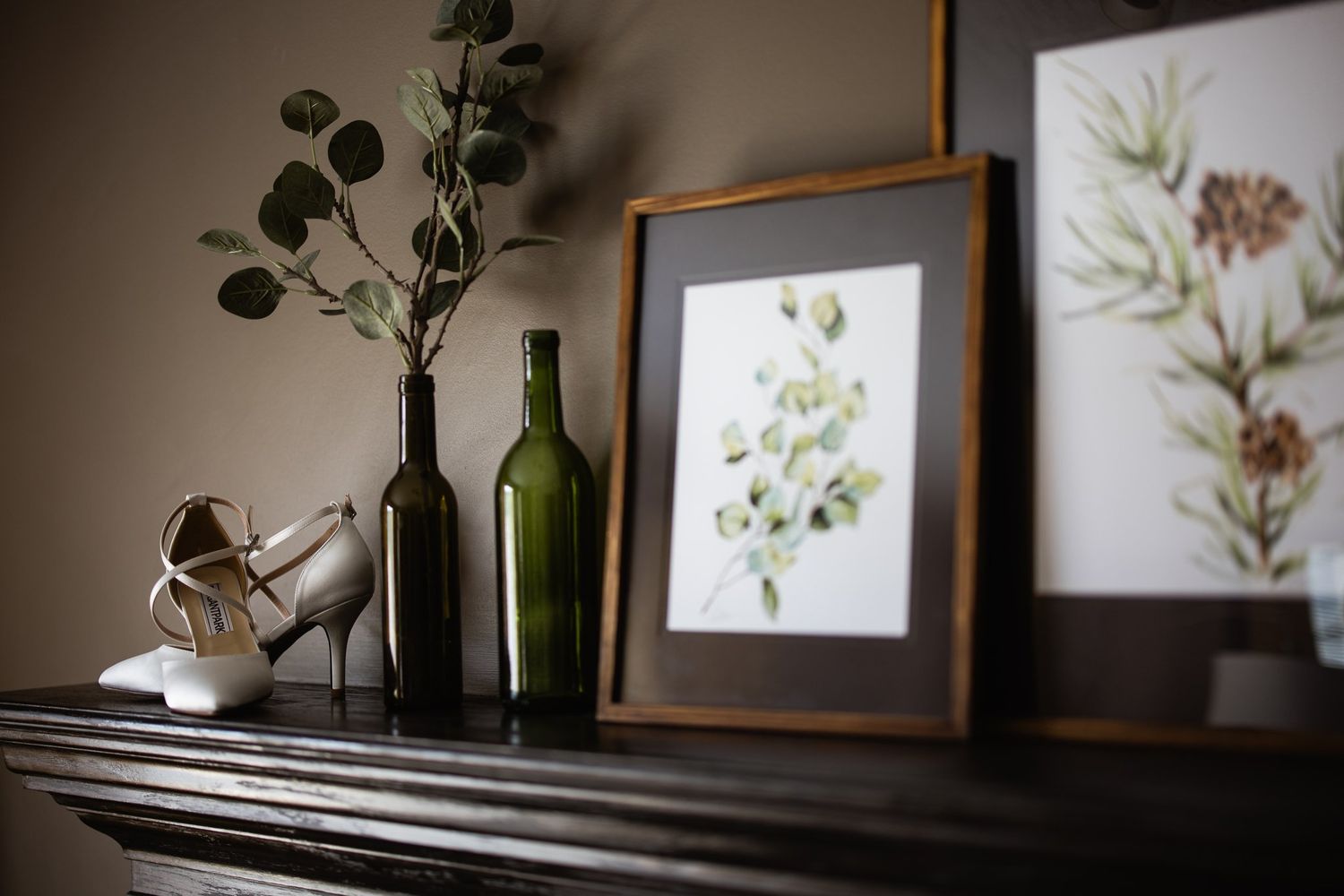 Bridal heels on a dark mantel beside a bottle vase and framed botanical prints