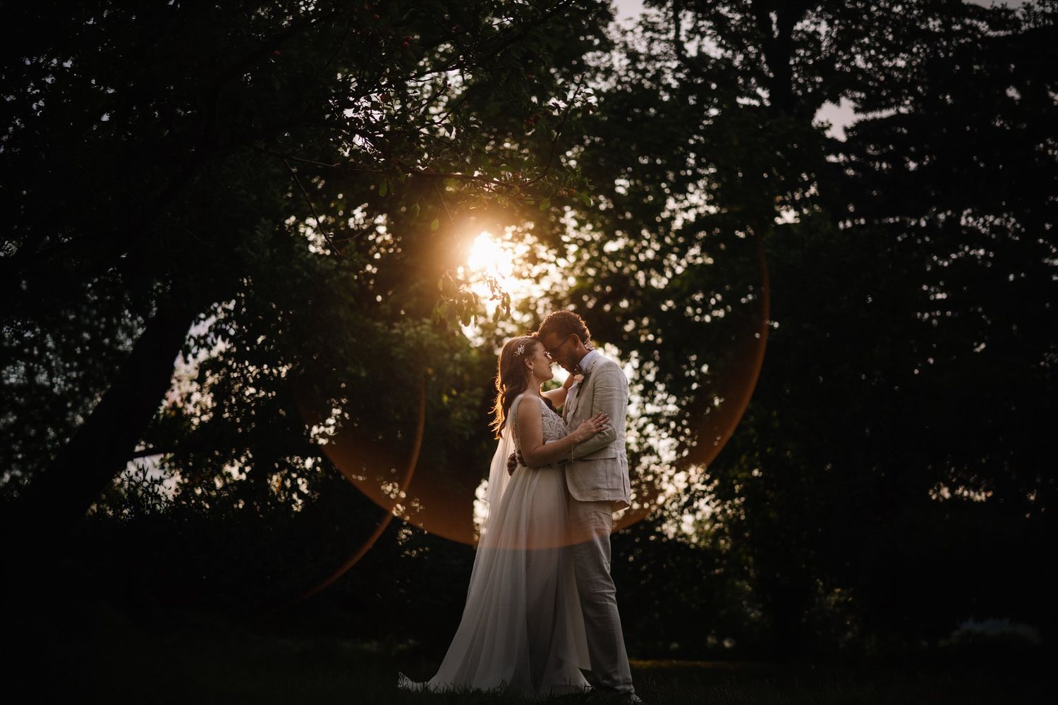 Couple kissing in dark trees with golden sun flare bursting behind them