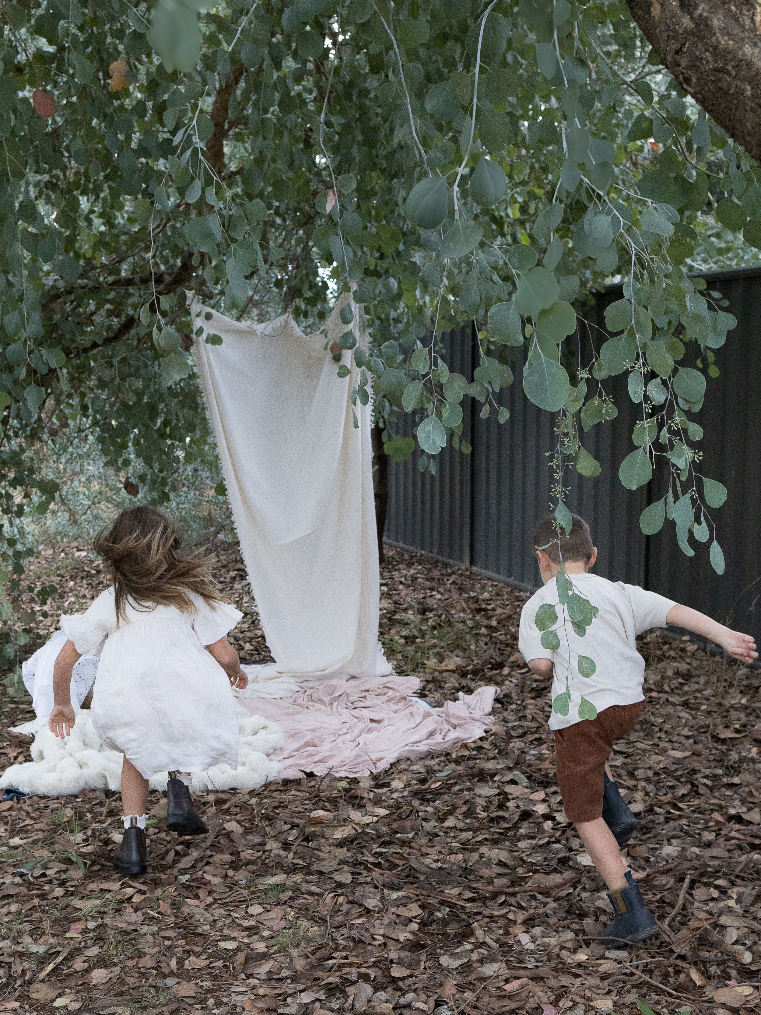Two children playing outdoors under eucalyptus tree with white fabric hanging, surrounded by fallen leaves.