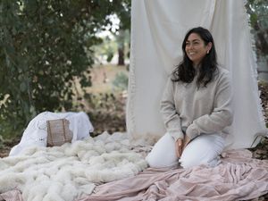 Woman in gray sweater and white pants sits smiling outdoors on pink fabric near fluffy white wool.