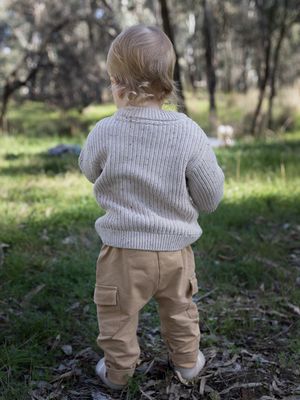 Toddler in beige knit sweater and tan cargo pants standing outdoors in a natural bush setting.