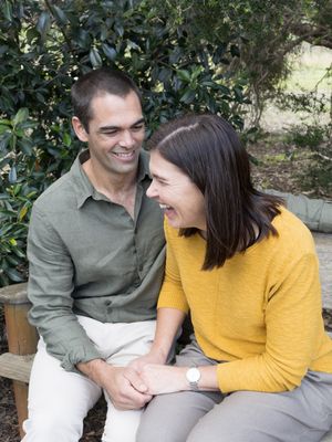 Happy couple sitting on a wooden bench outdoors, laughing together, man in green shirt, woman in yellow sweater.