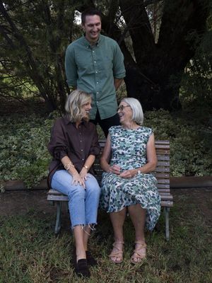 Three people posing outdoors on a wooden bench surrounded by lush greenery in a relaxed family portrait.