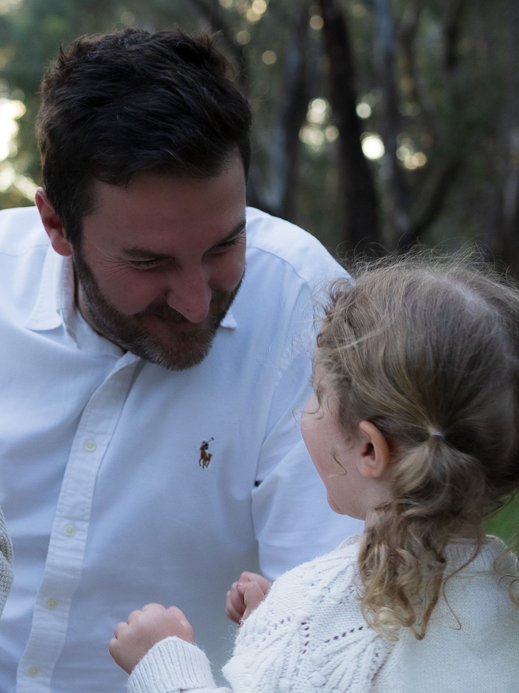 A bearded man in a white polo shirt smiles warmly at a young blonde girl in a white sweater outdoors.